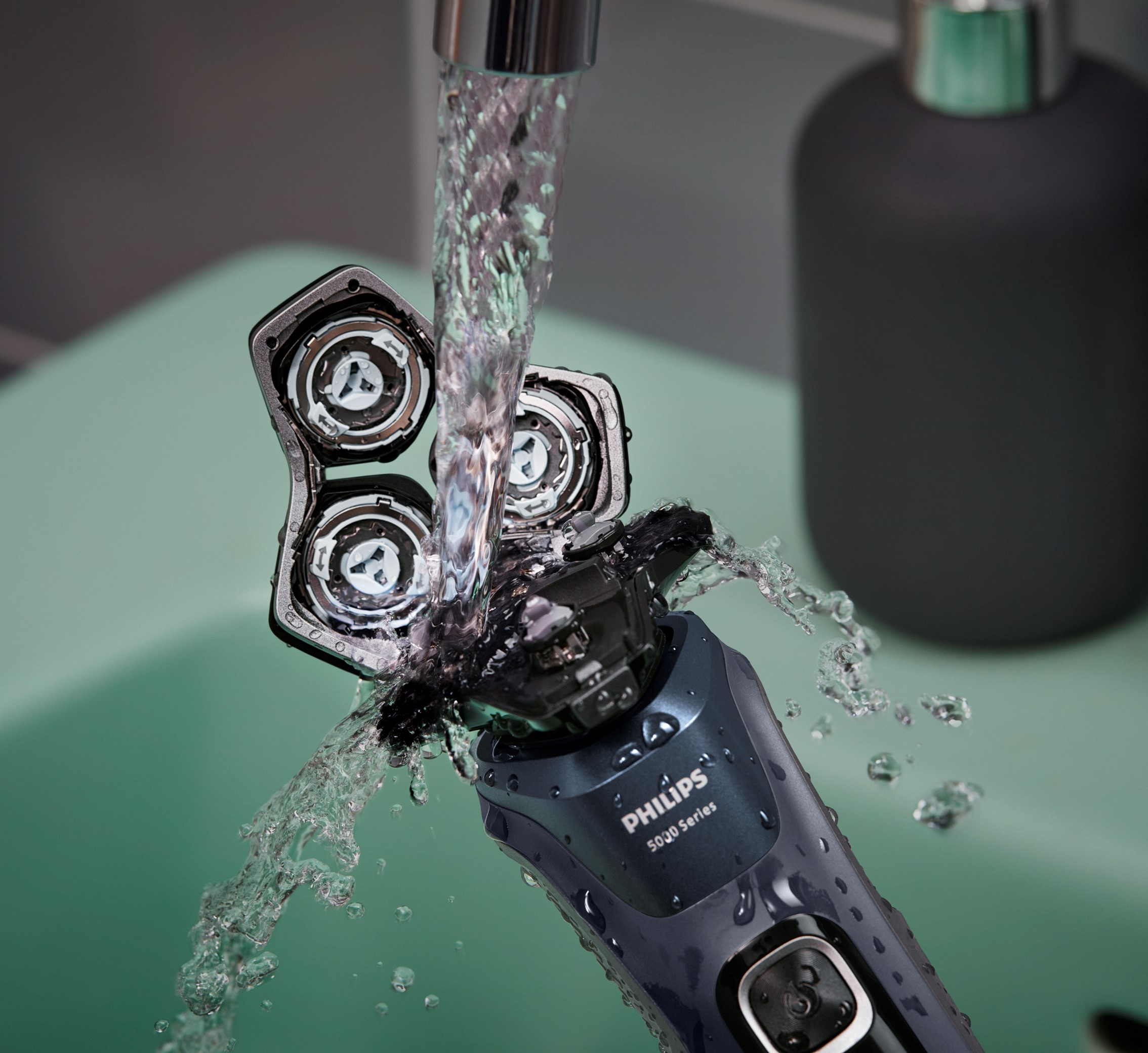 Cabezal de afeitadora eléctrica enjuagándose con agua sobre un lavamanos verde.