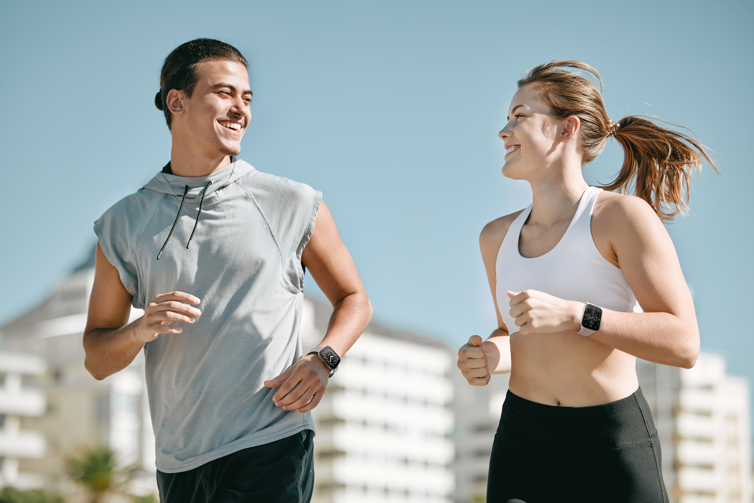 Un hombre y una mujer corren juntos al aire libre, usando relojes inteligentes y sonriendo el uno al otro.