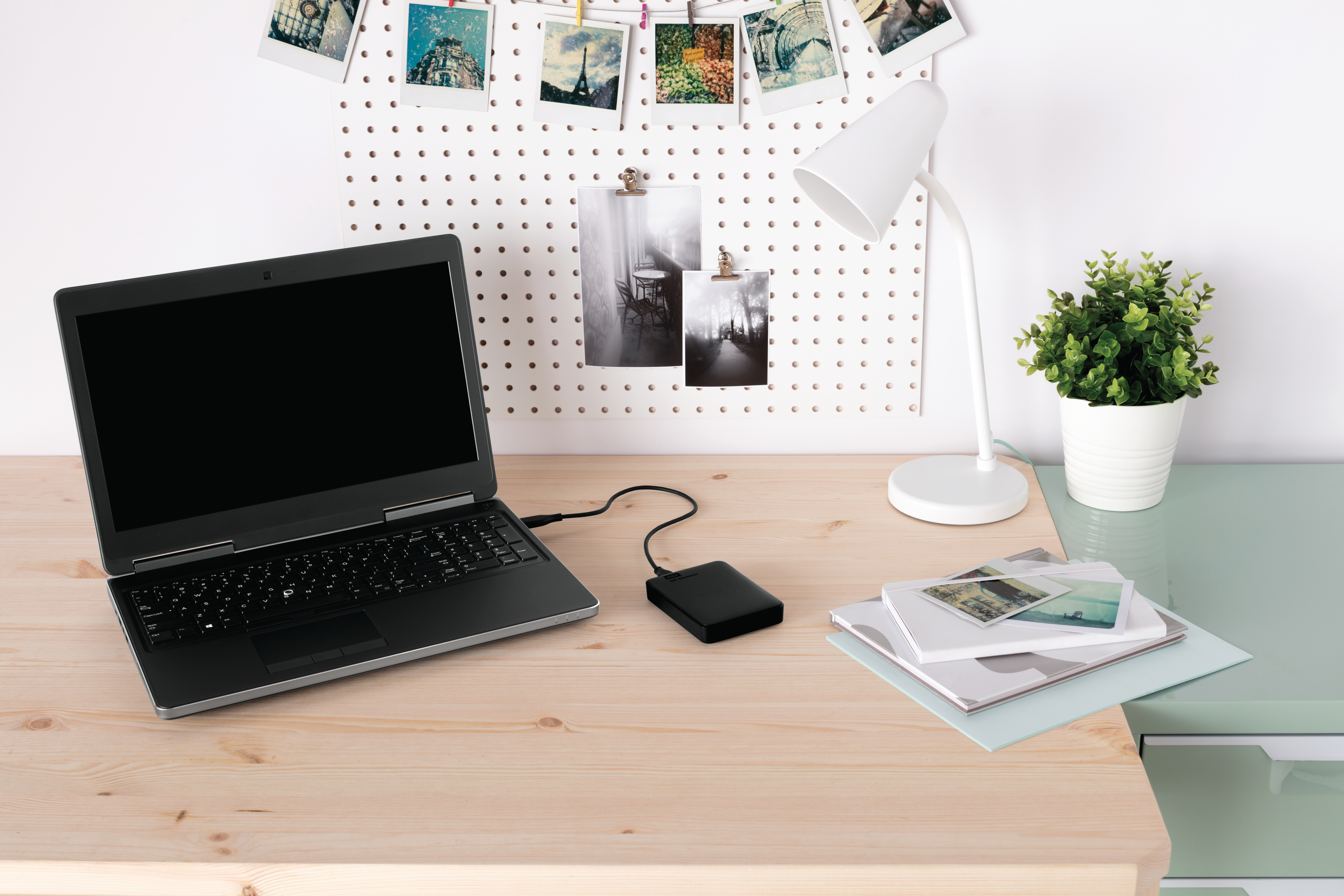 Laptop, Lampe und Pflanzen auf einem Holzschreibtisch. Fotos an einer weißen Lochwand im Hintergrund.