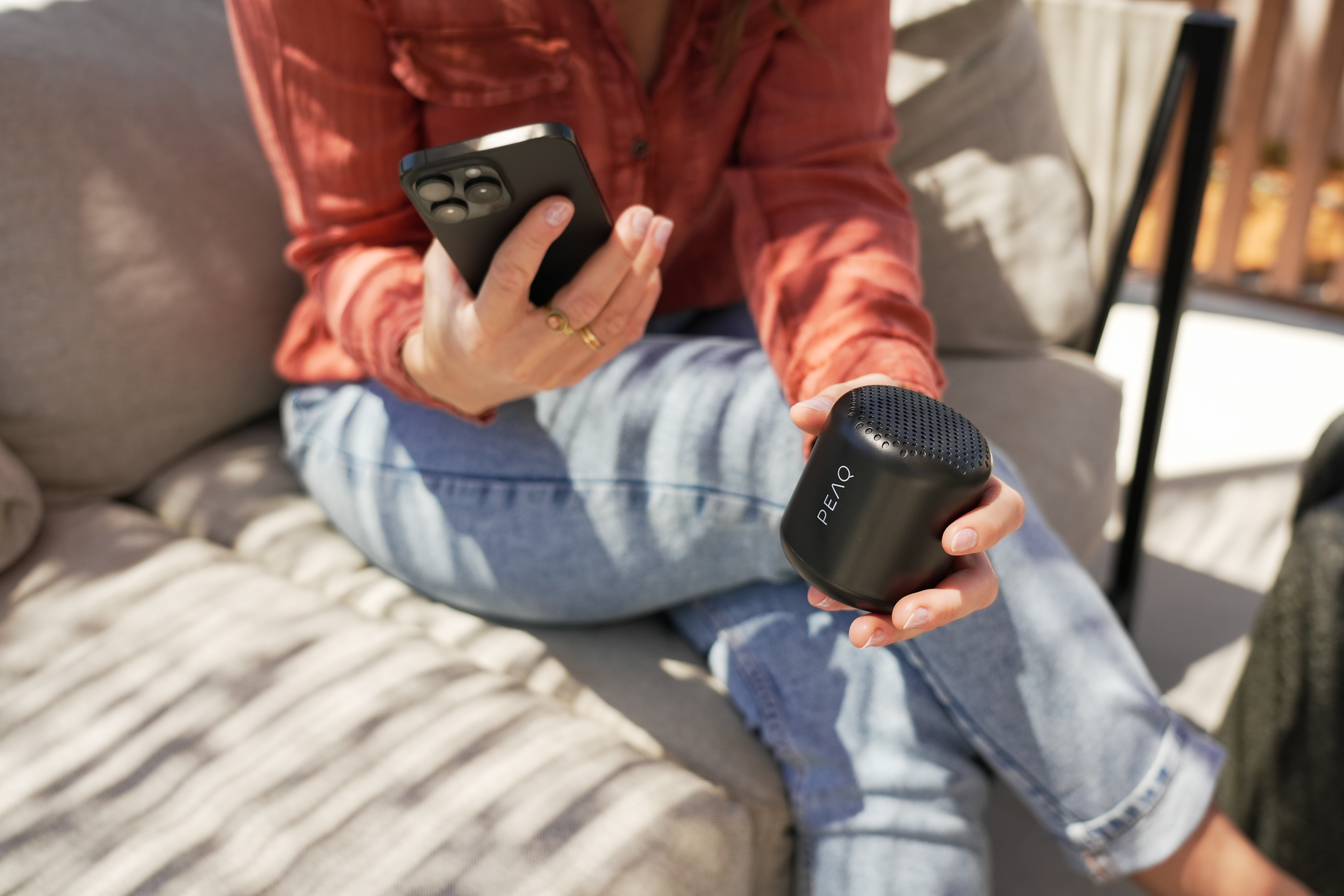 Frau hält einen schwarzen Lautsprecher und ein Smartphone. Sie sitzt auf einem Sofa.