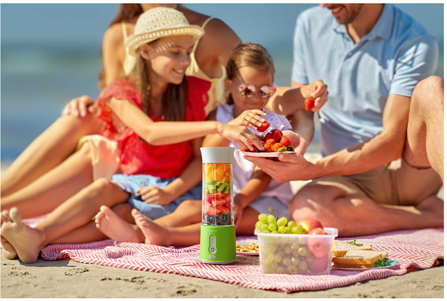 Család a strandon, gyümölcsöt eszik egy turmixgépből. Piknik takaró és tenger.