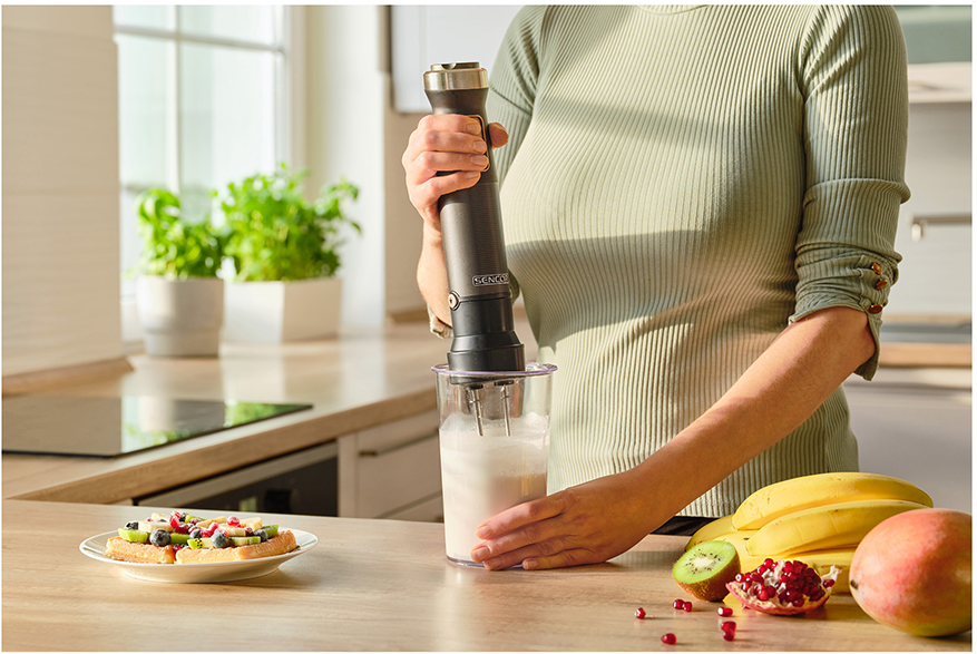 Személy kézi mixert használ a konyhában. Smoothie üvegben, gyümölcsök és szendvics.