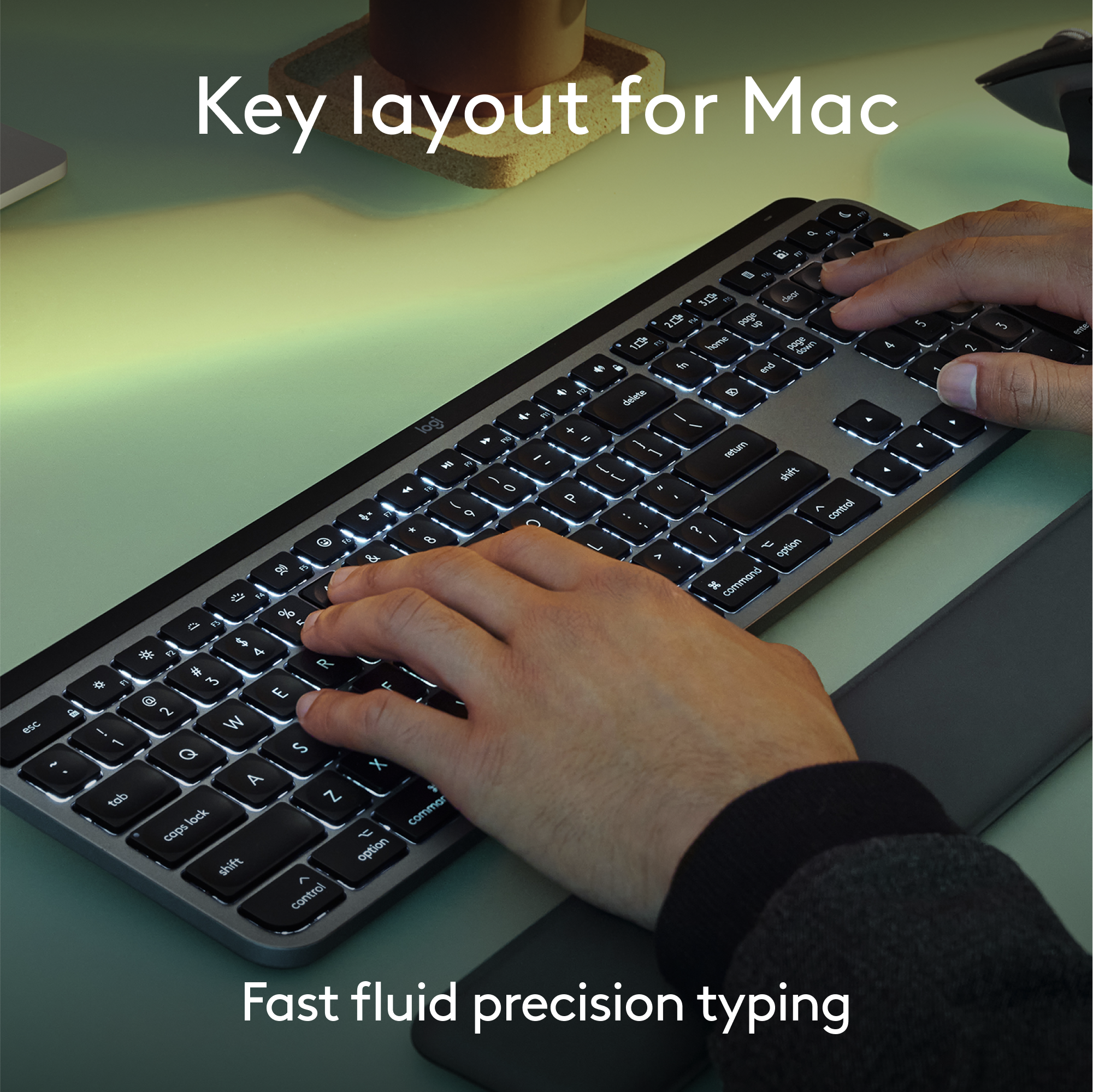 Mani che digitano su una tastiera retroilluminata, con layout Mac, e un poggiapolsi su una superficie verde.