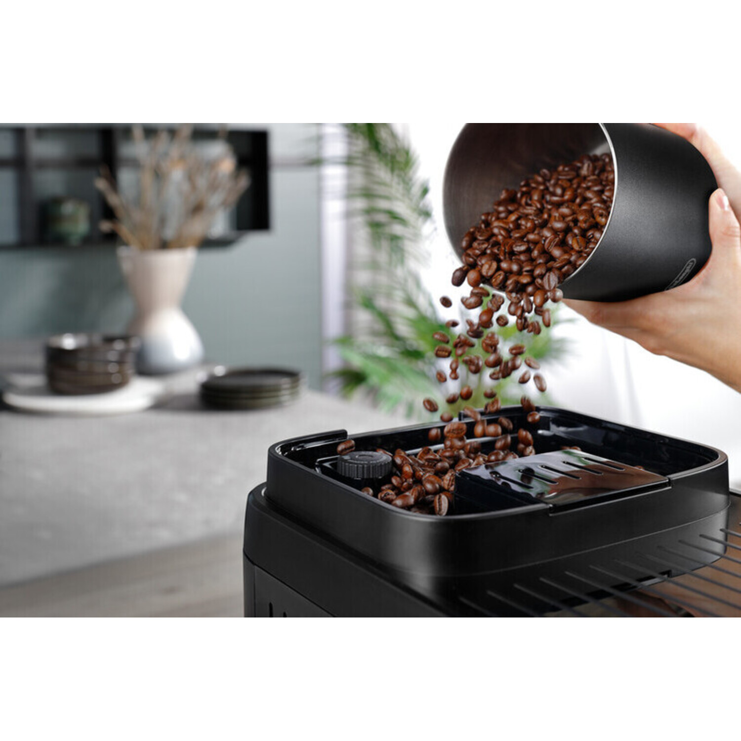 Coffee beans pouring into a black coffee machine, a person's hand holding the container.
