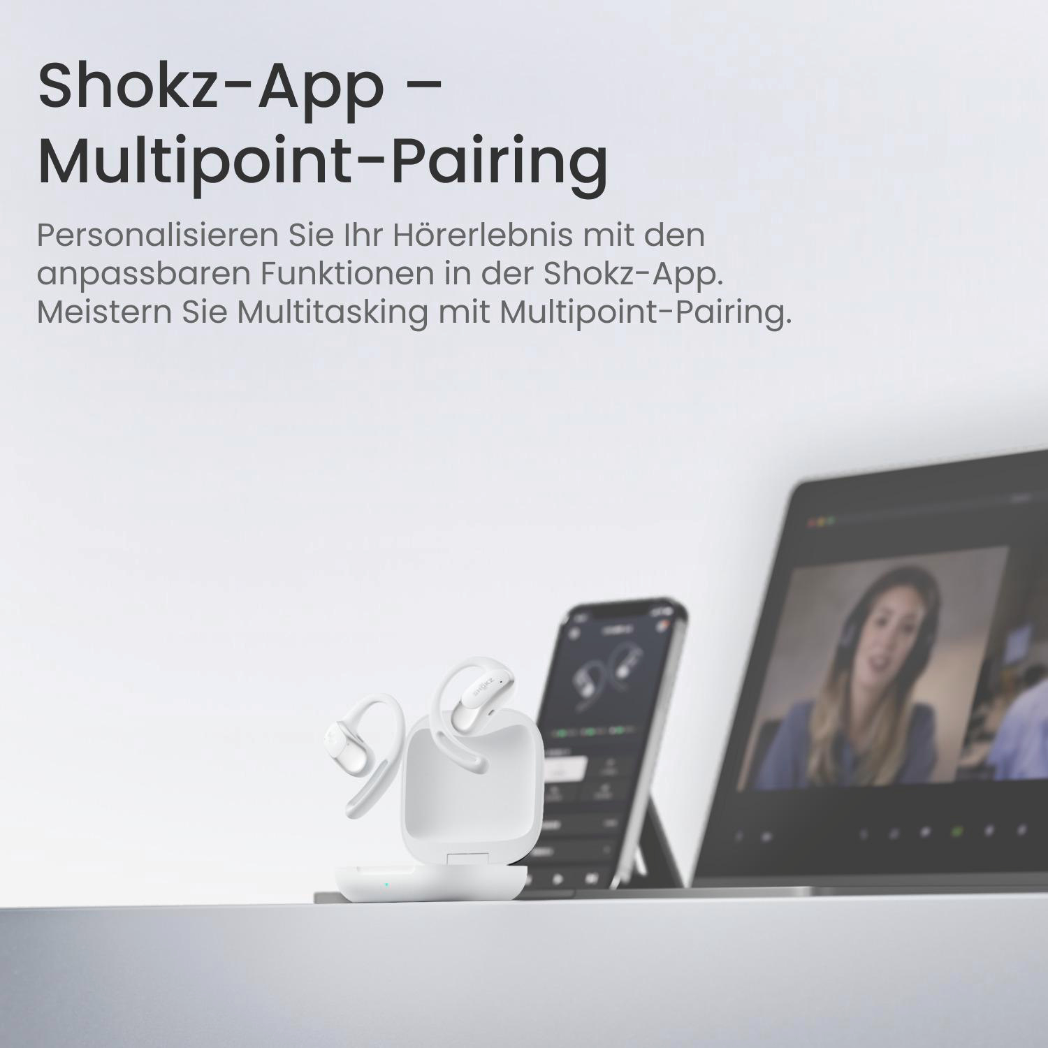Weiße Shokz-Ohrhörer in der Ladebox, Telefon mit App-Anzeige und Laptop mit Videoanruf, alles auf weißer Oberfläche.