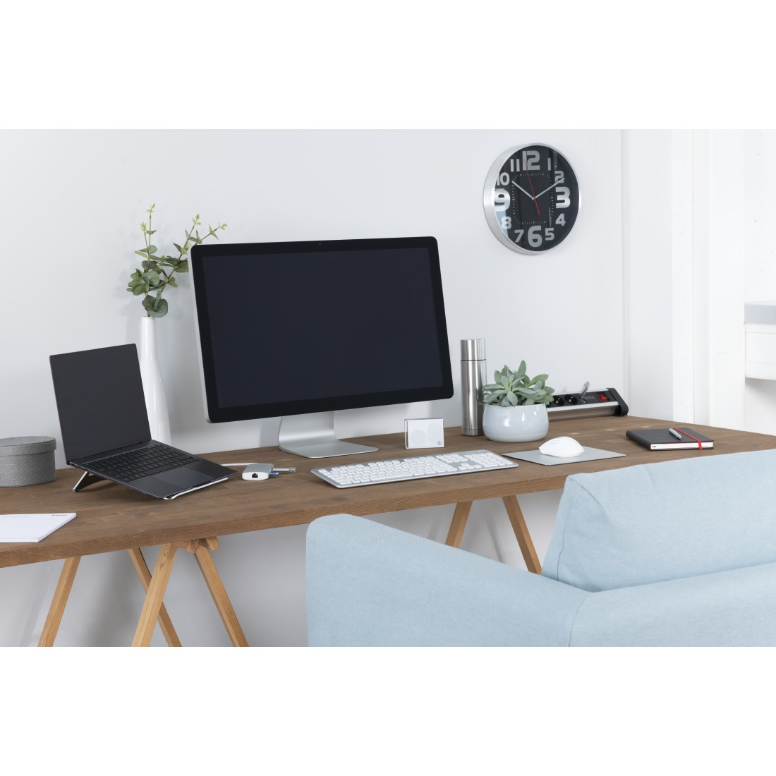 Espacio de trabajo con portátil, monitor, teclado, ratón, reloj y plantas. Escritorio de madera con sofá azul y pared blanca.