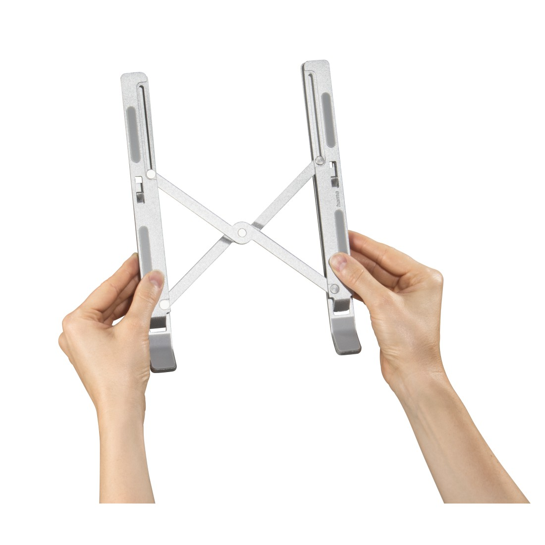 Manos sosteniendo un soporte para portátil plateado. El soporte está plegado en forma de X, fondo blanco.