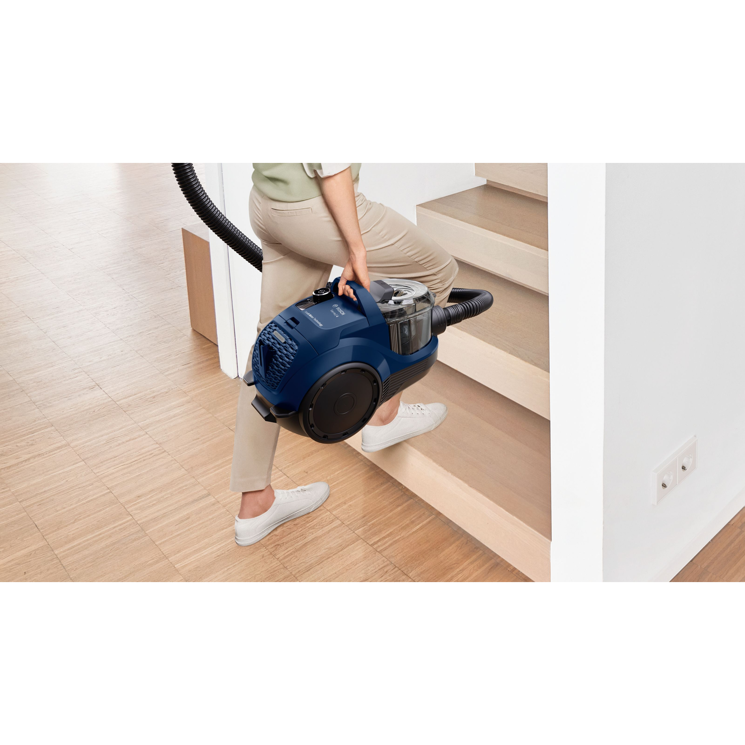 A person carrying a blue vacuum cleaner up wooden stairs. White wall with a light switch in the background.