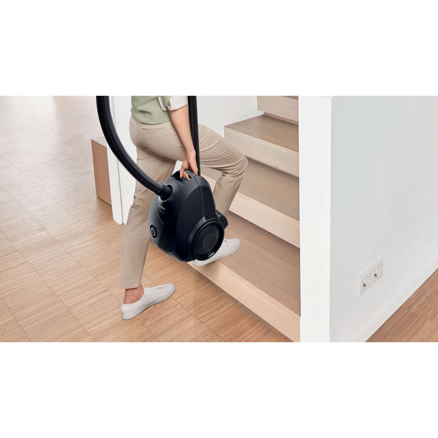 A person carrying a black vacuum cleaner up wooden stairs, towards a white wall.