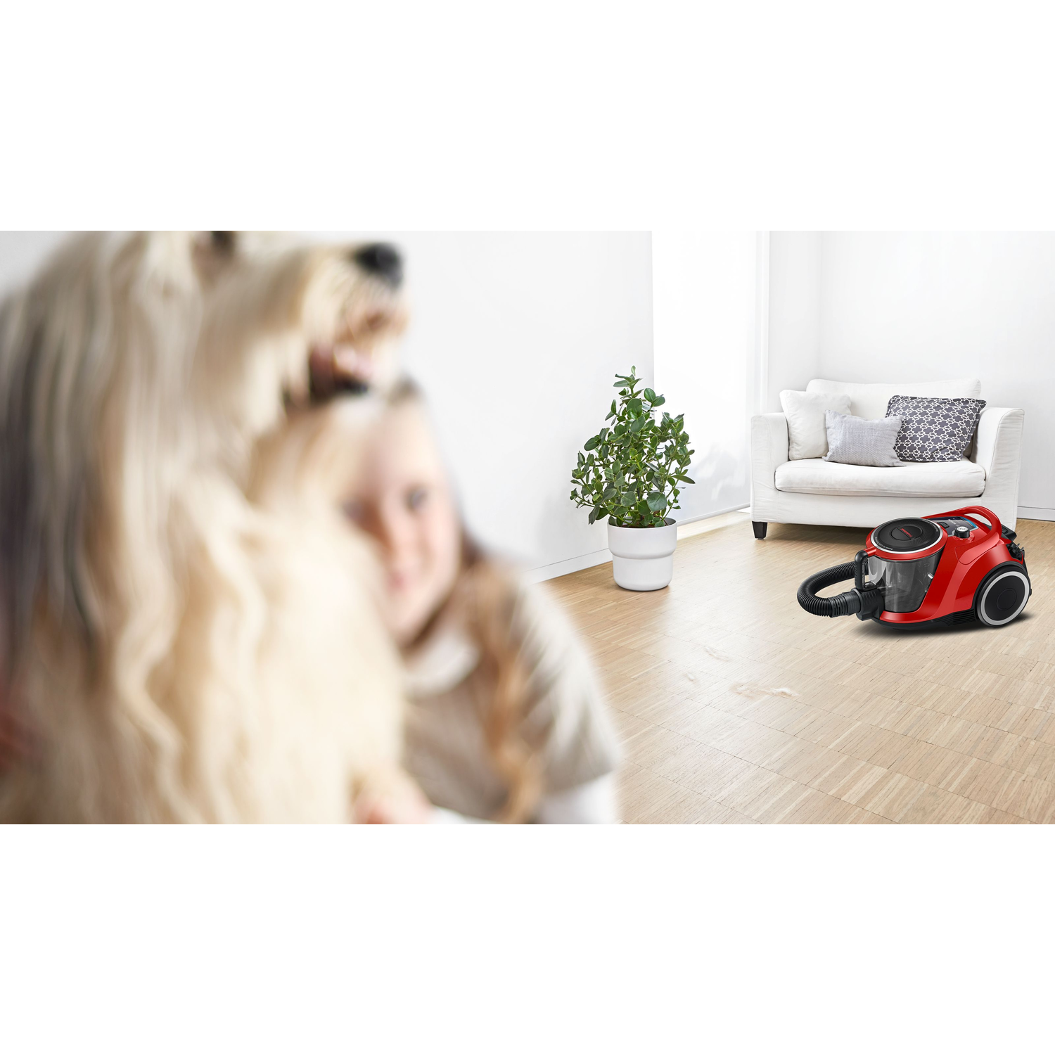 A girl is hugging a dog, the vacuum cleaner is in the background. White walls and wooden floor.