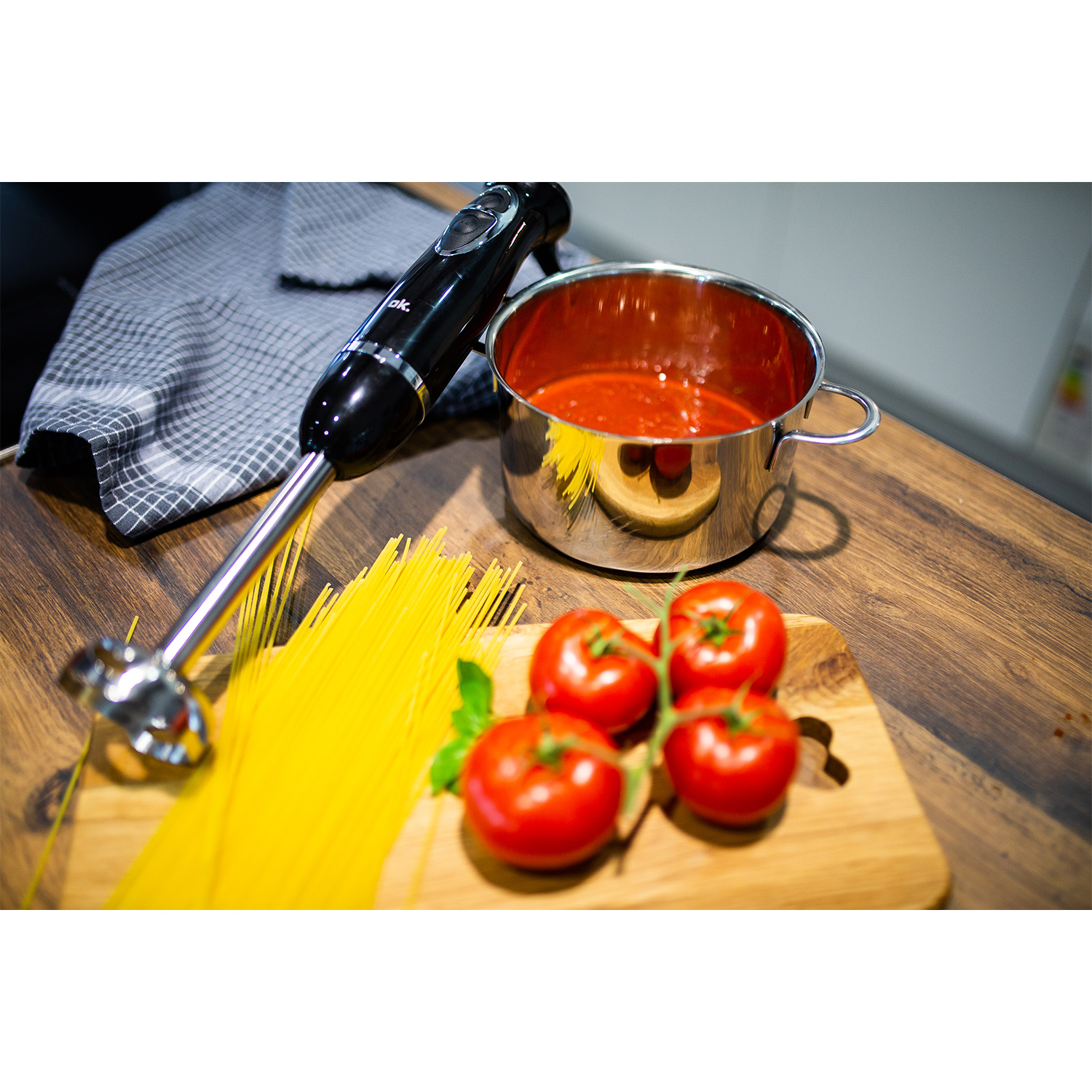 Frullatore a immersione nero, pentola di salsa di pomodoro, pomodori e pasta su un tavolo di legno.