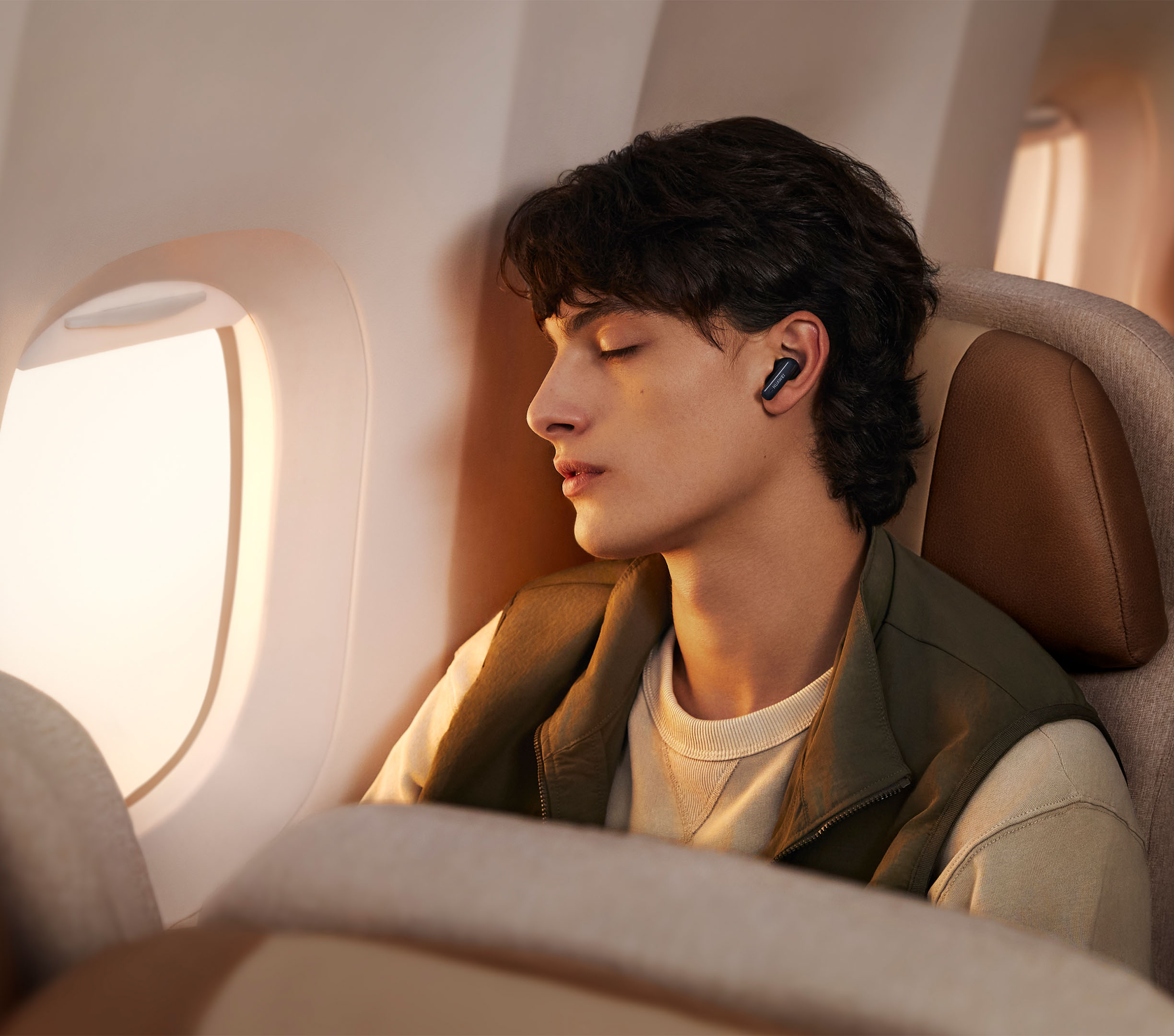 Młody mężczyzna śpiący w samolocie. Nosi słuchawki i kurtkę. Widoczne jest okno.