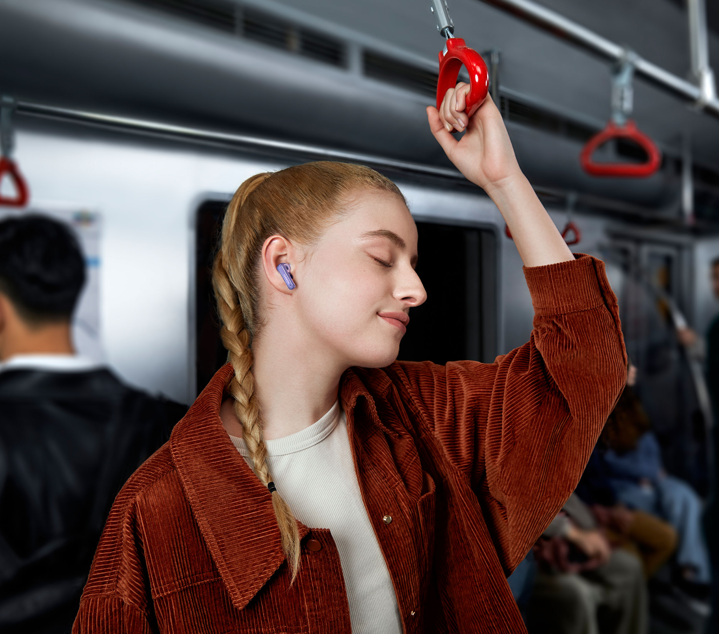 Kobieta w brązowej sztruksowej kurtce w pociągu. Ma na sobie fioletową słuchawkę i trzyma się poręczy.