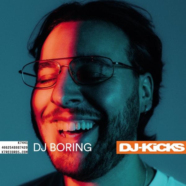 DJ Boring lächelt, trägt eine Brille, mit blau-rotem Licht im Hintergrund und dem Text DJ-Kicks.