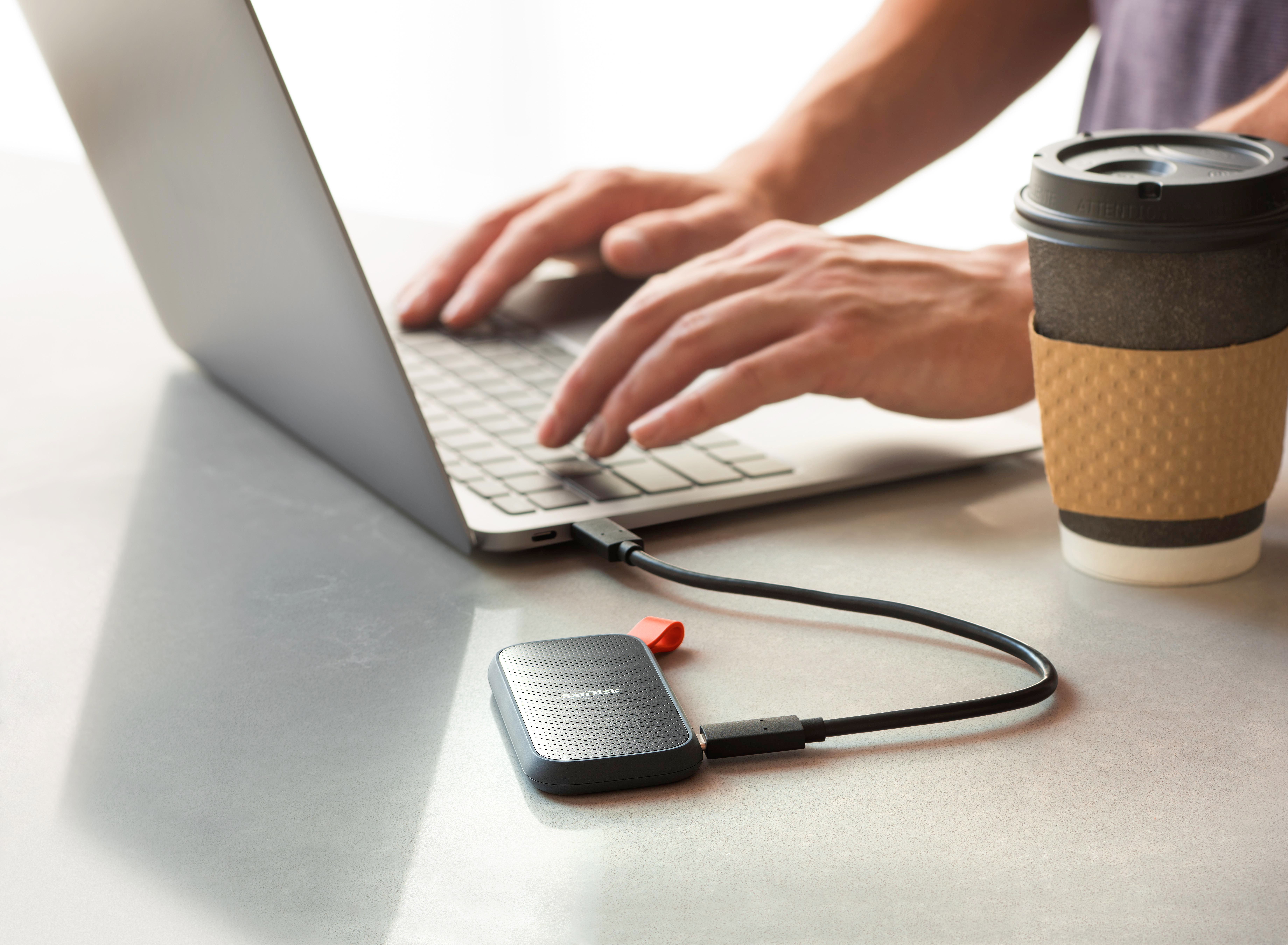 Person tippt auf Laptop, verbunden mit externer Festplatte und Kaffeetasse auf dem Tisch.