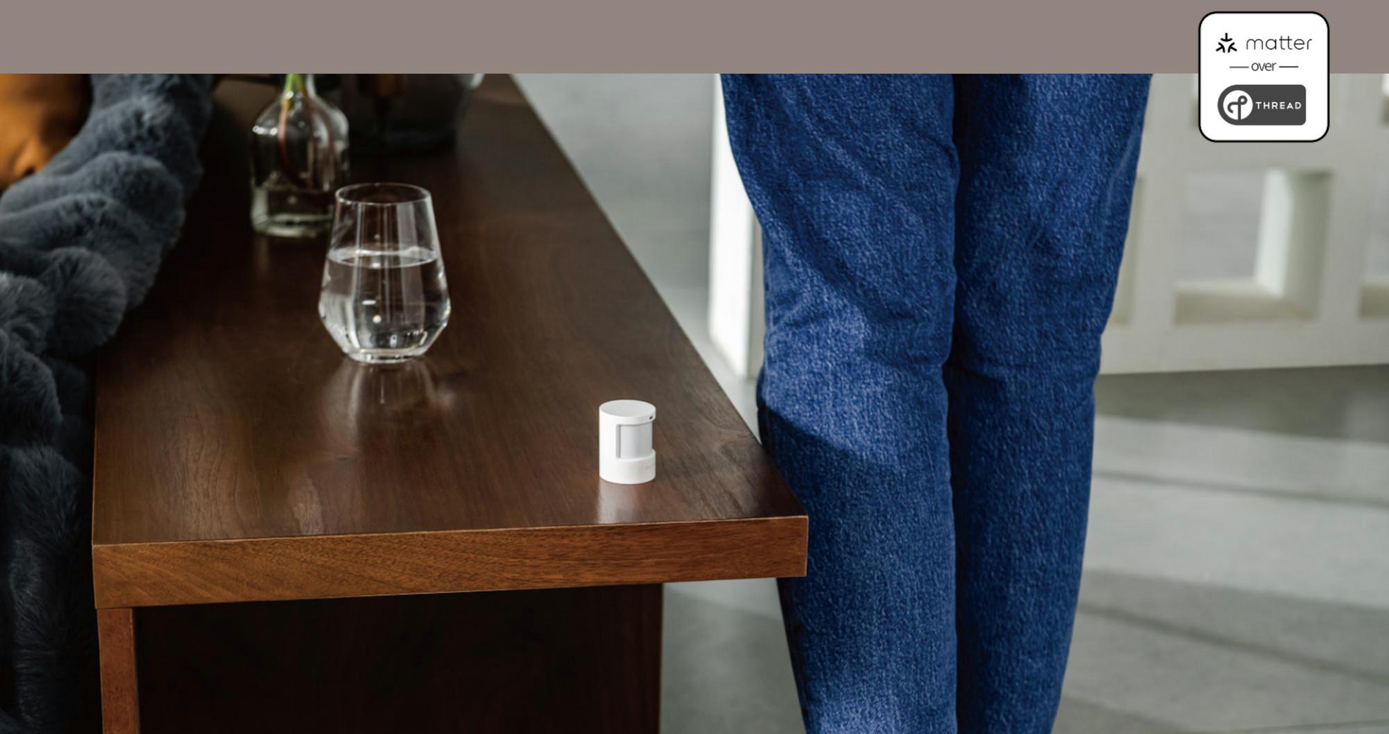 Ein weißer Sensor liegt auf einem Holztisch. Ein Glas Wasser und eine Person in Jeans sind sichtbar.
