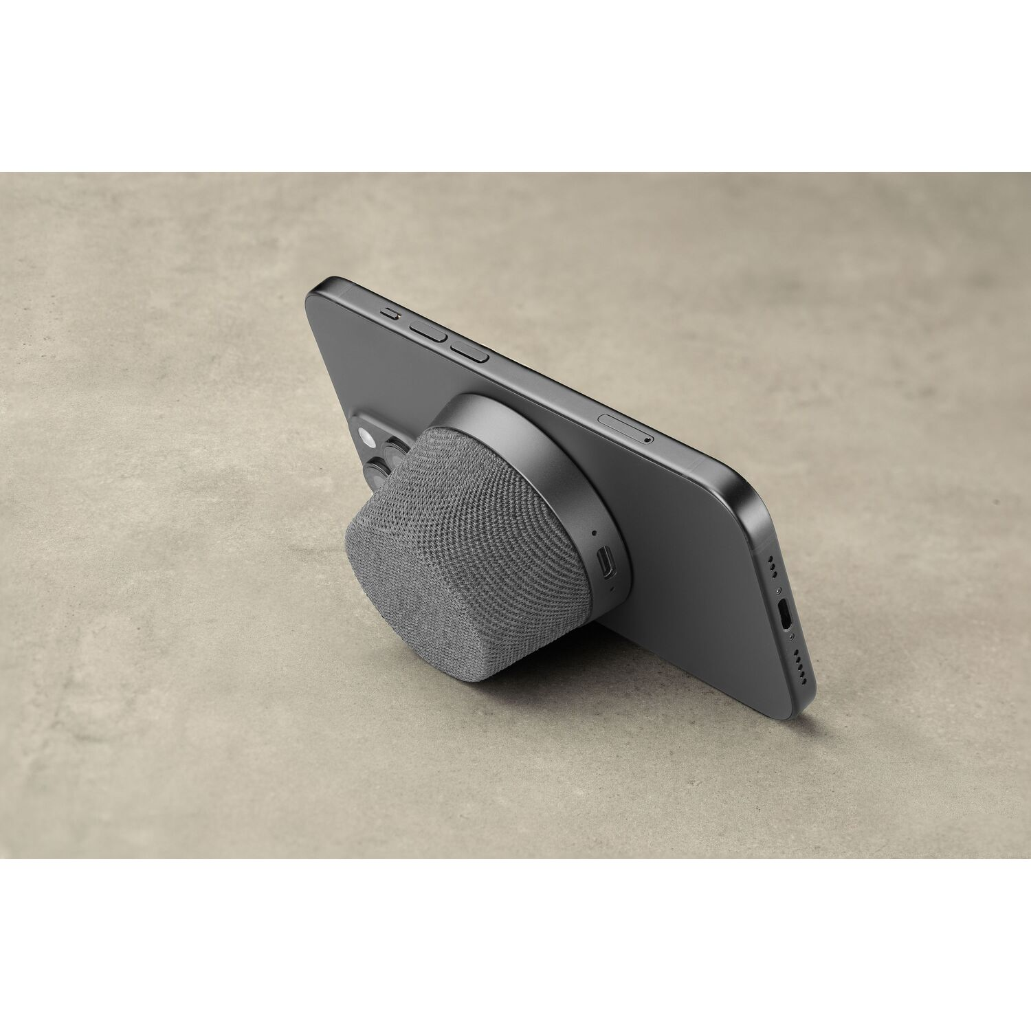 A dark grey phone with a grey fabric-covered accessory attached to the back, resting on a surface.