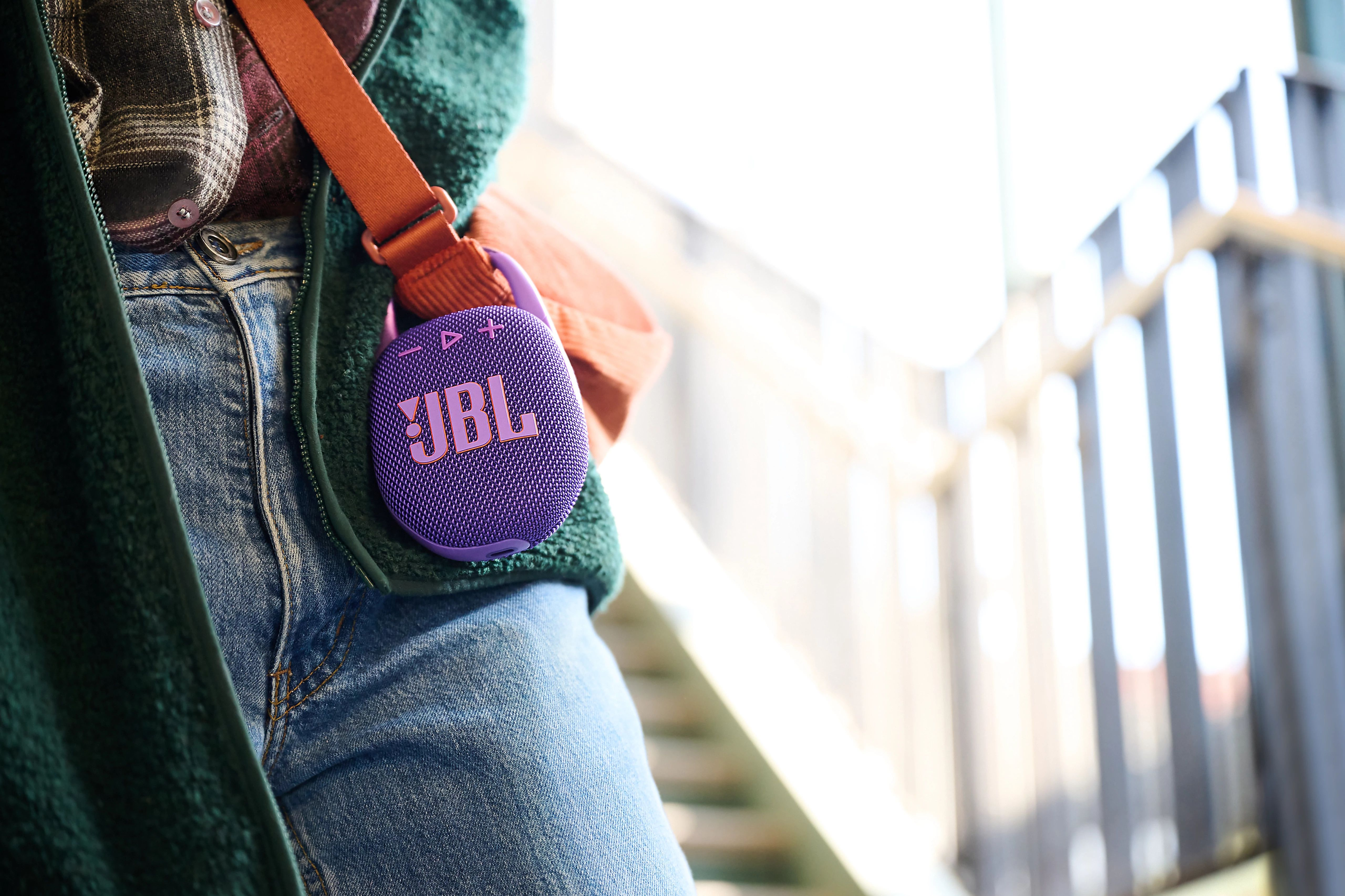 Bir kişinin kot pantolon cebine takılmış mor ve pembe bir JBL hoparlör, turuncu bir kayış ile.