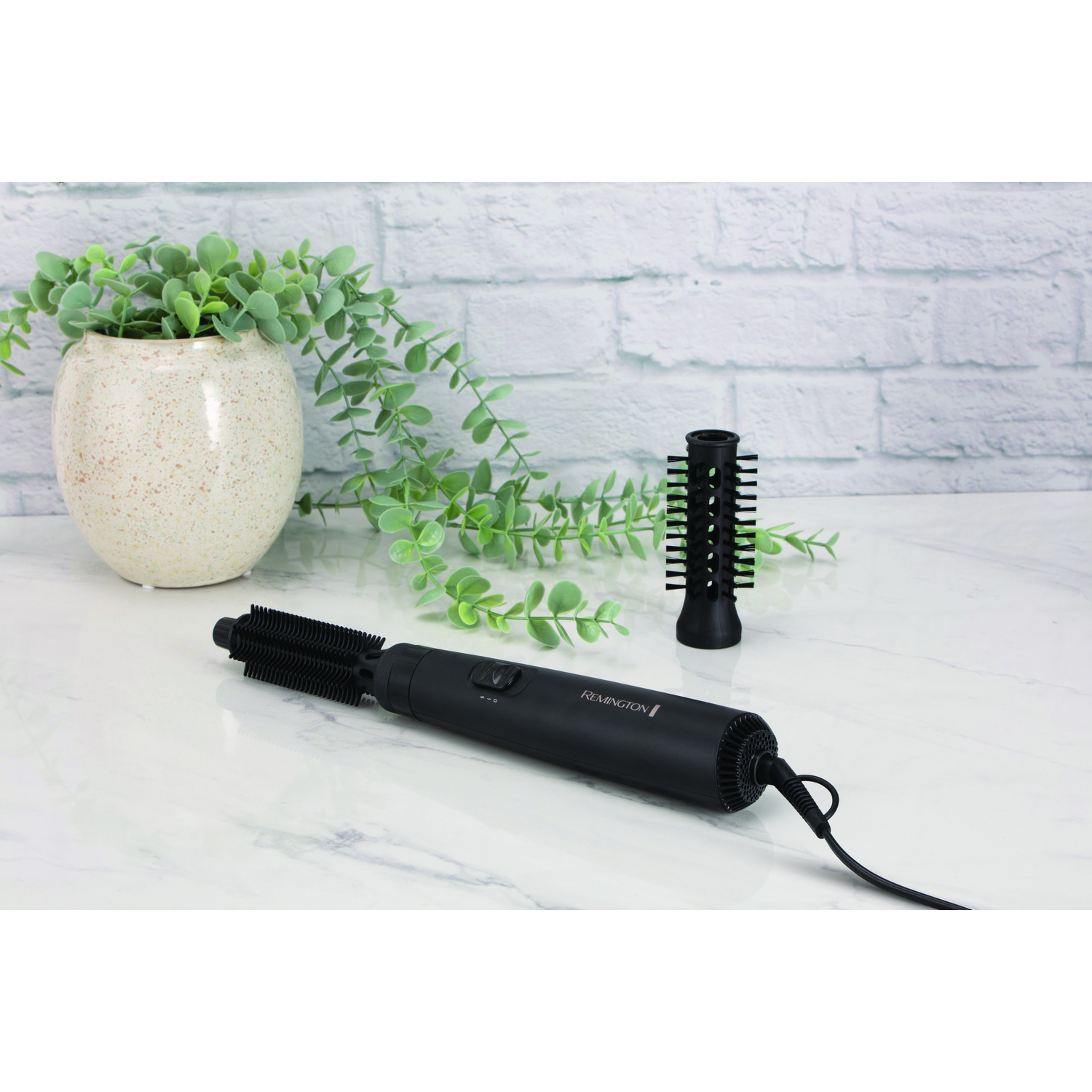 Styliseur de cheveux noir avec deux accessoires brosse. Une plante en pot à gauche.