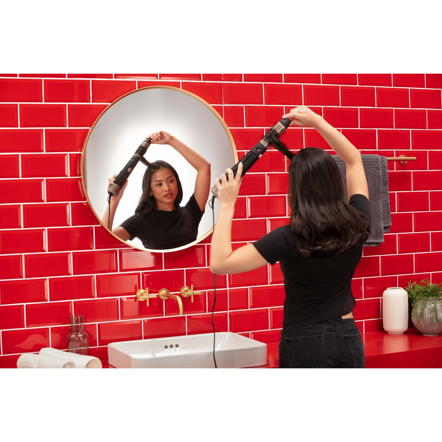 Femme se séchant les cheveux dans une salle de bain carrelée de rouge, se regarde dans un miroir rond. Robinet doré et lavabo blanc.