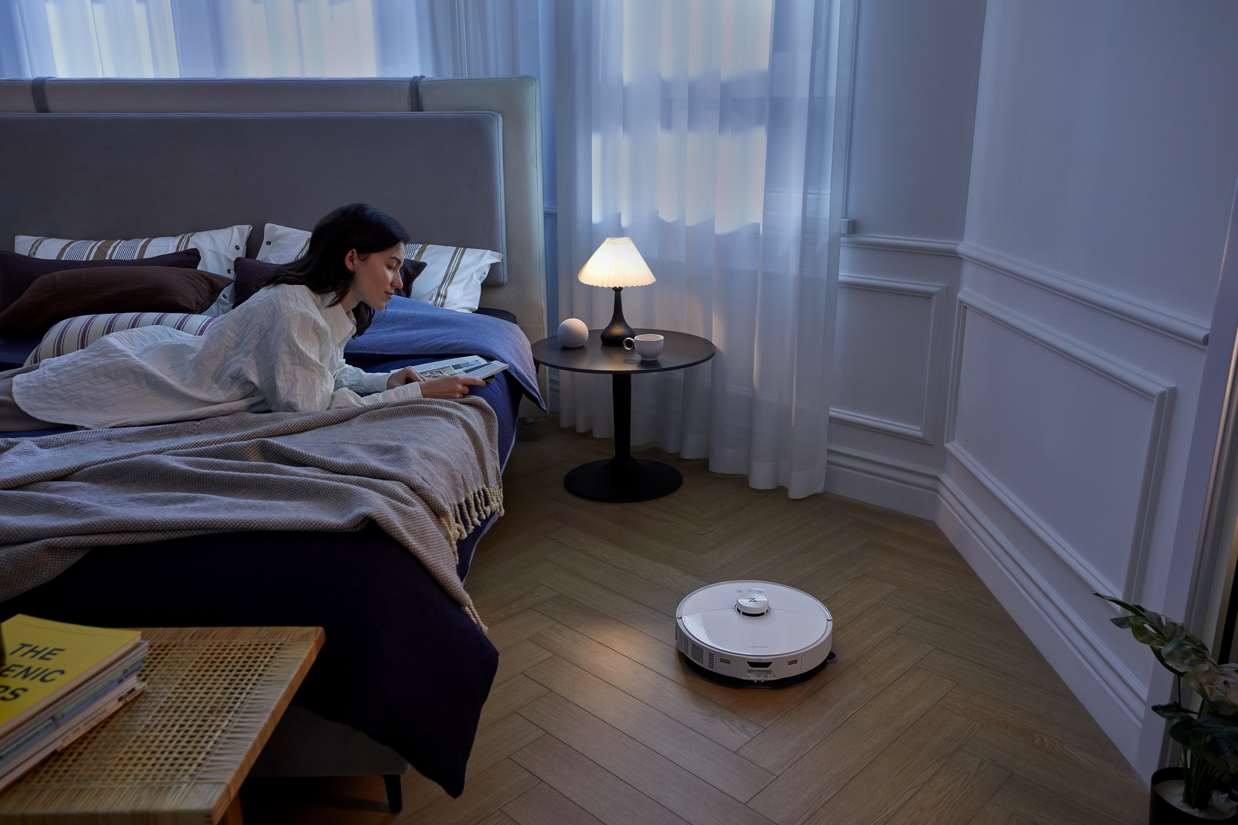 Kobieta czyta na łóżku z lampą i stolikiem. Robot sprzątający na drewnianej podłodze.