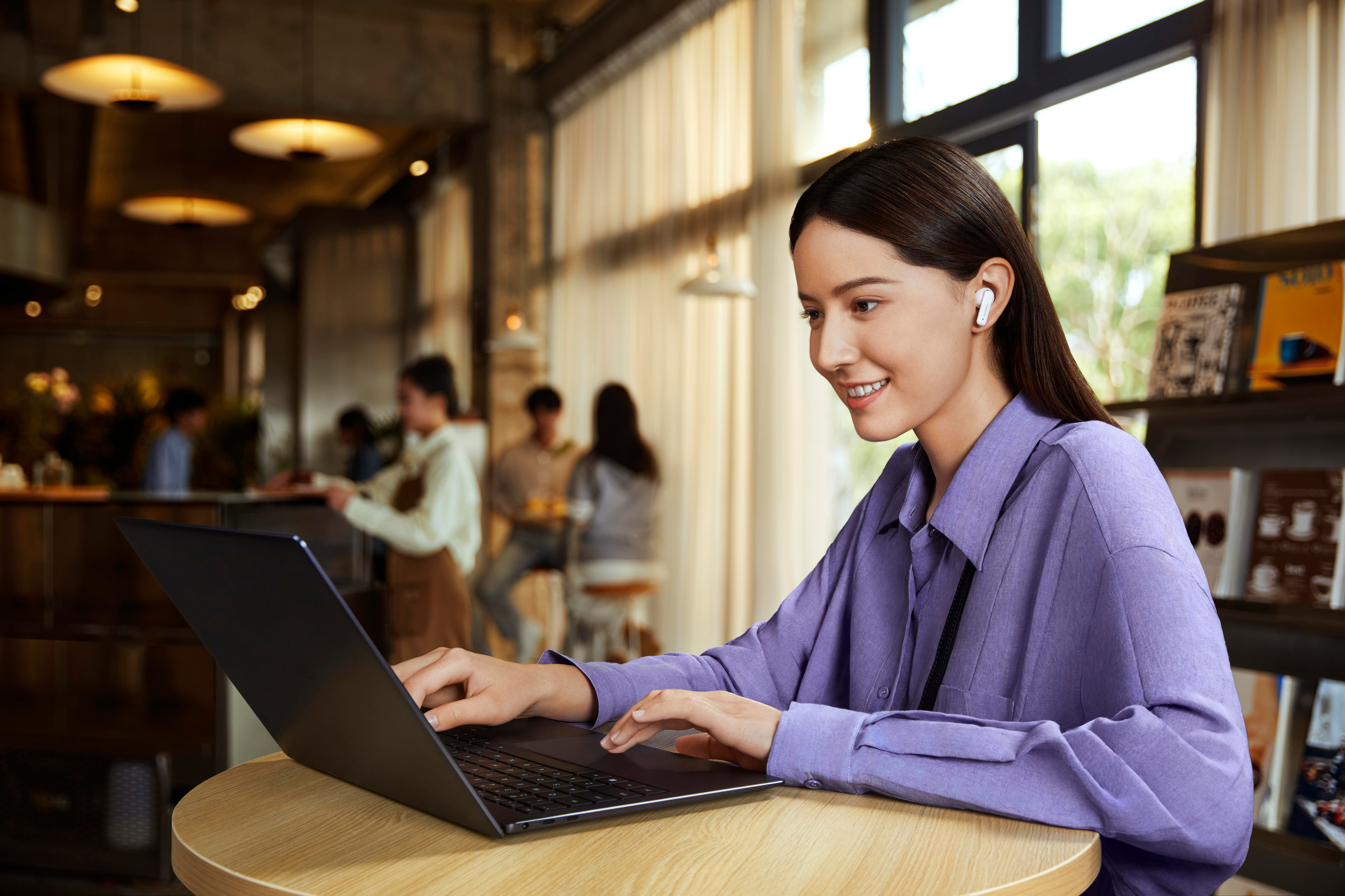 Eine Frau in einem lila Hemd benutzt einen Laptop und Ohrhörer in einem Café. Bücher und andere Personen im Hintergrund.