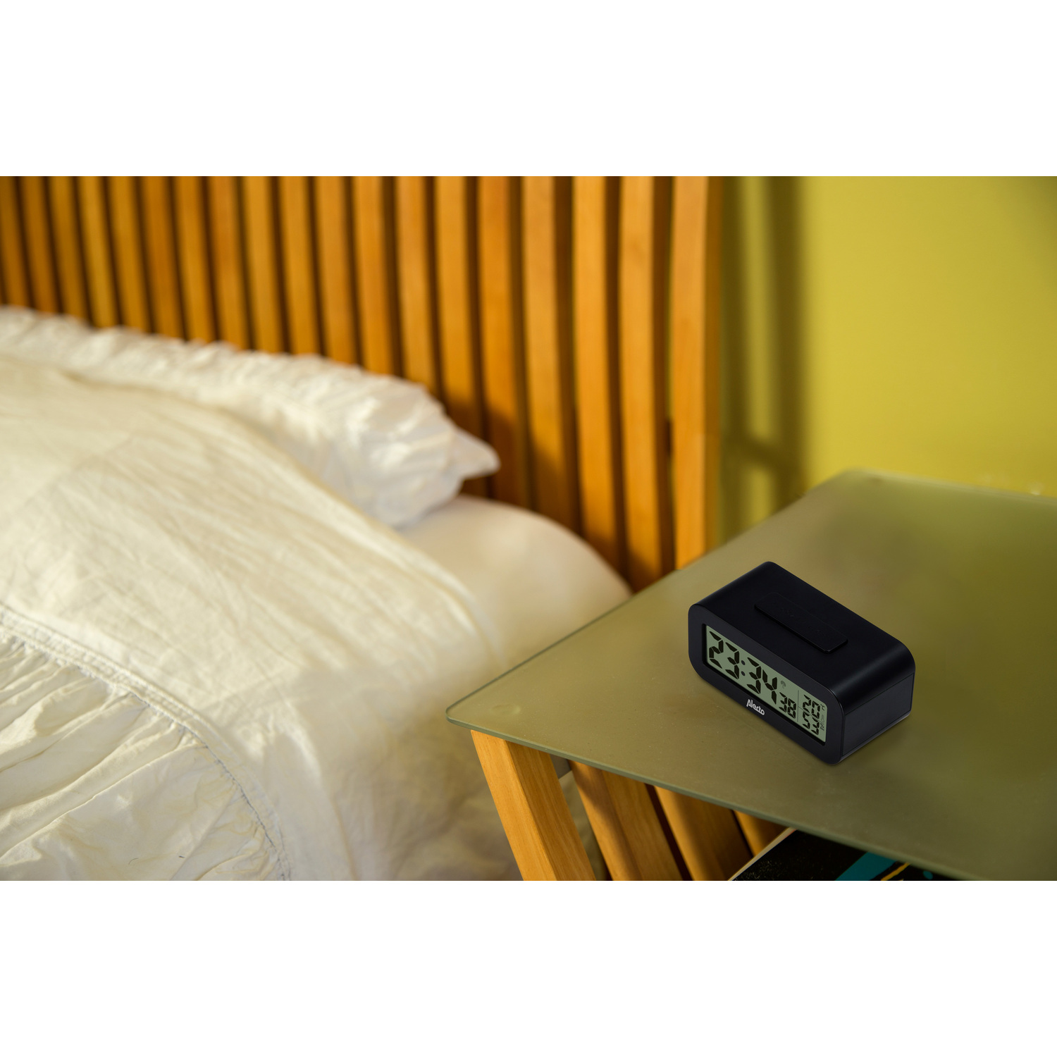 Un réveil numérique noir sur une table à côté d'un lit. L'horloge affiche l'heure.