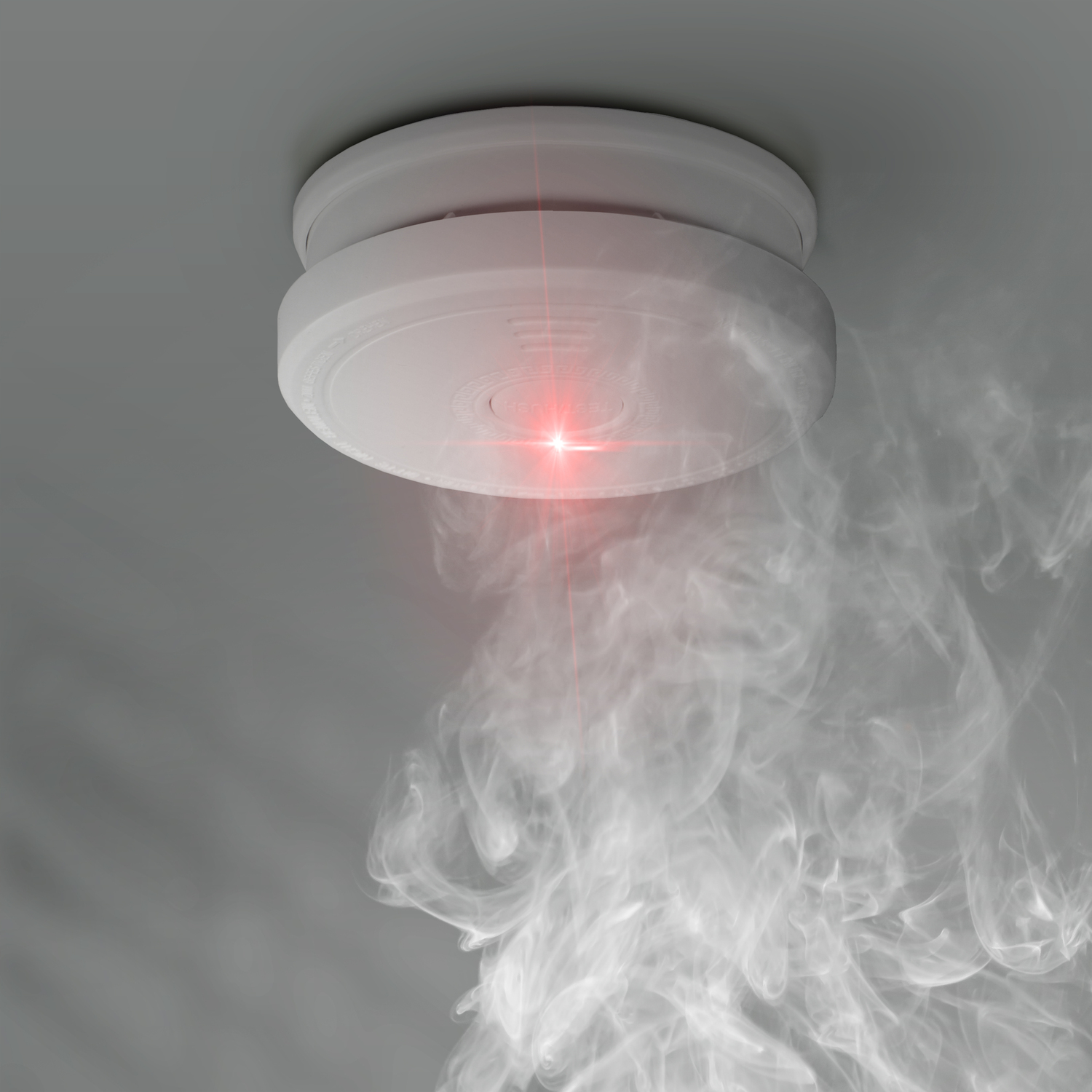 Smoke detector mounted on a gray ceiling, emitting smoke and a red light.