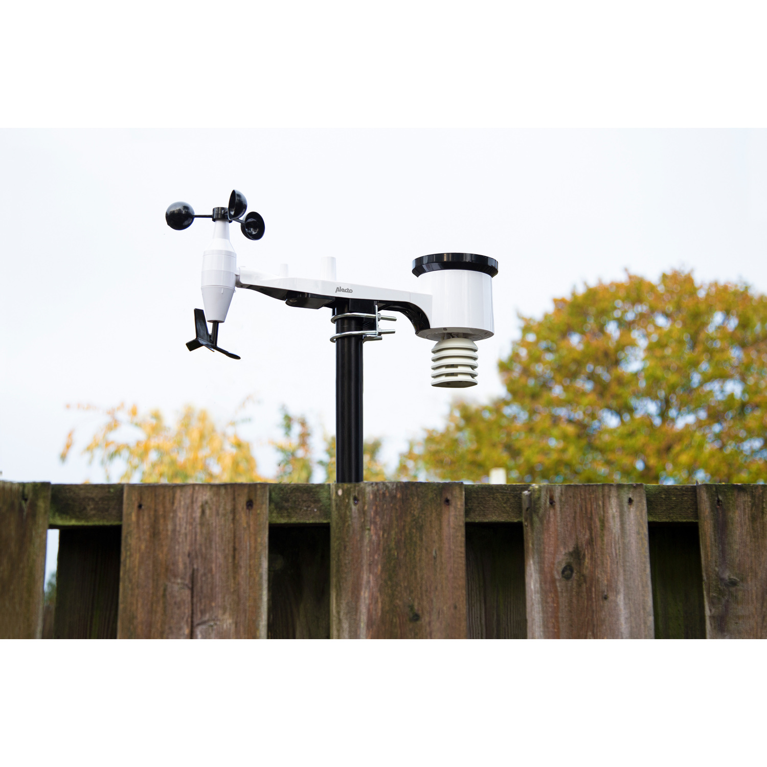 A white weather station on a black pole.  Wooden fence and trees in the background.