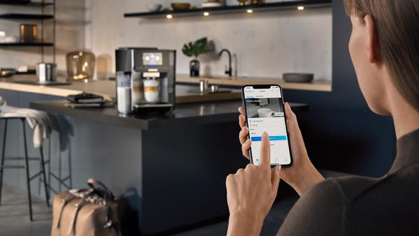 Eine Frau, die ein Telefon benutzt, um eine Kaffeemaschine in einer Küche zu steuern.