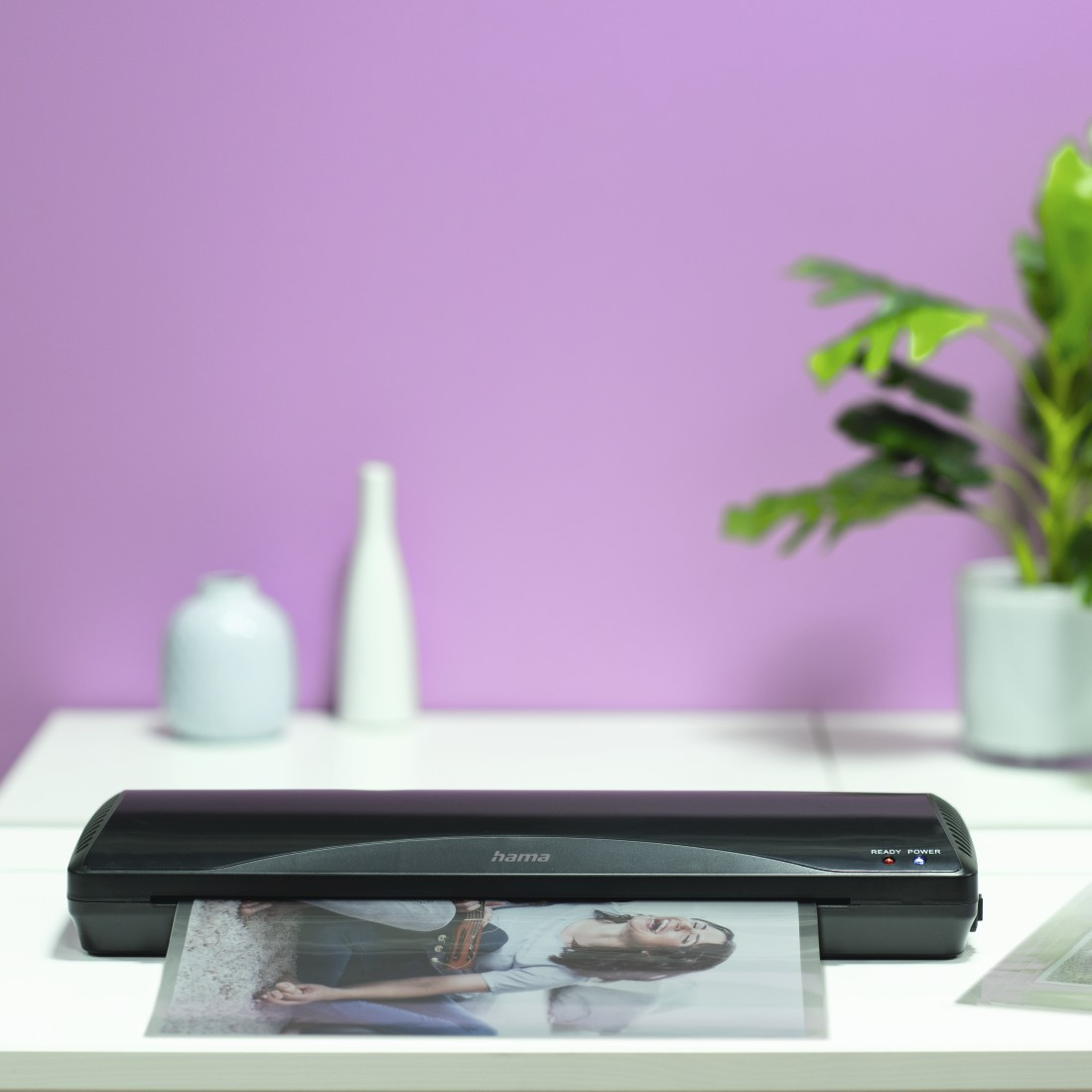 Laminadora con foto, planta y florero en mesa blanca contra pared rosa.