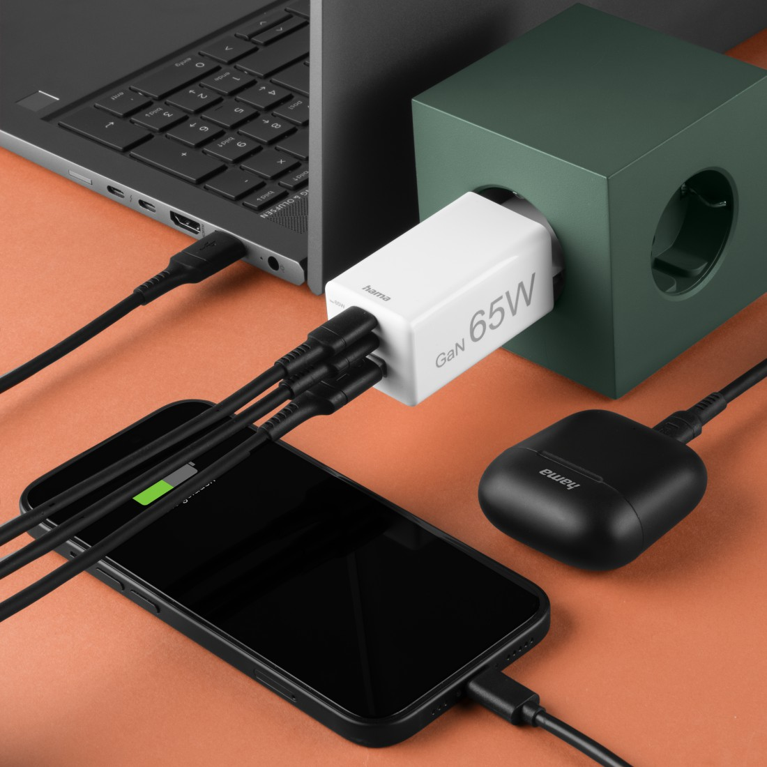 Un teléfono, una computadora portátil y unos auriculares se cargan con un cargador GaN de 65W sobre una superficie naranja.