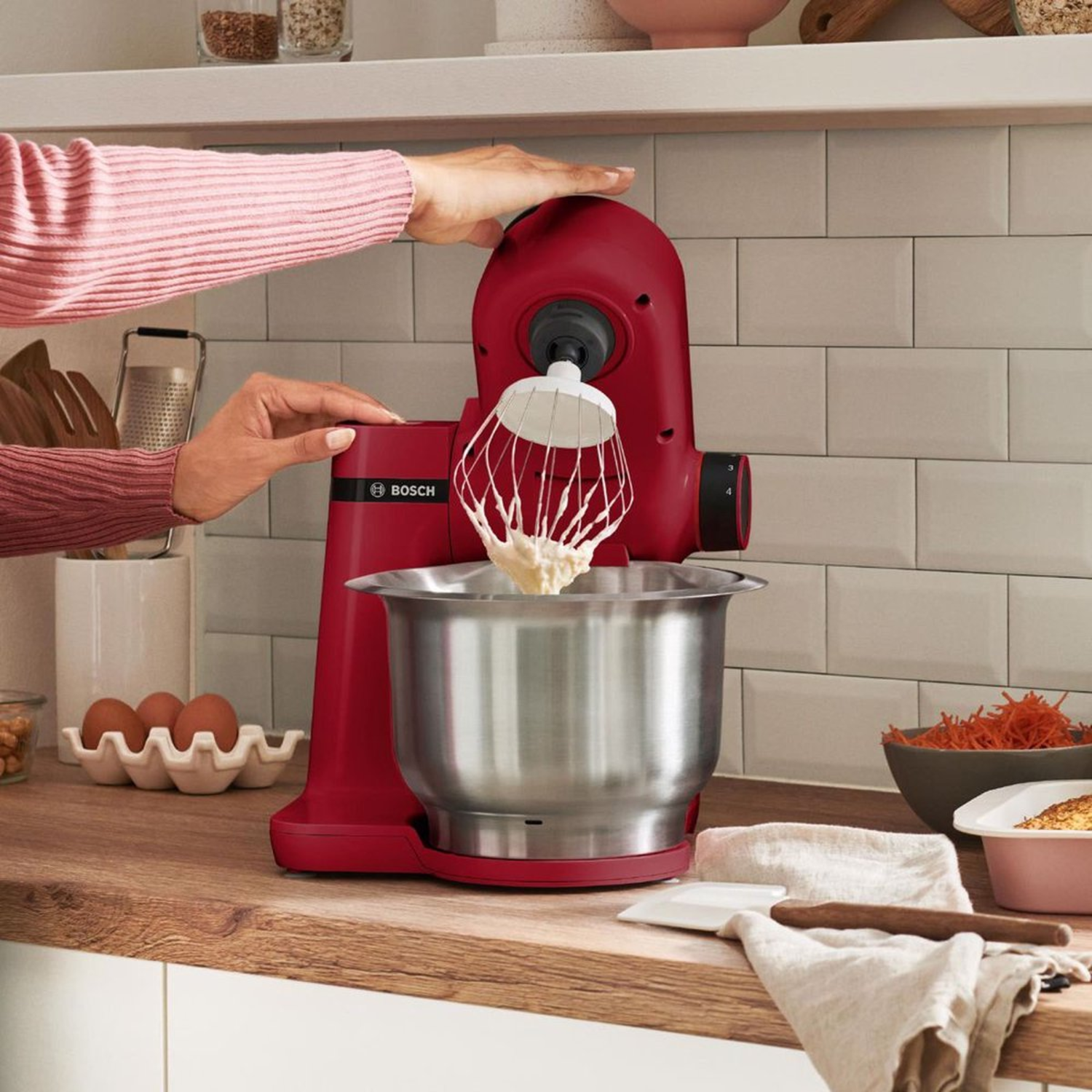 Een rode Bosch keukenmixer met een garde-opzetstuk die ingrediënten mengt in een zilveren kom, handen bedienen hem. Keukenomgeving.