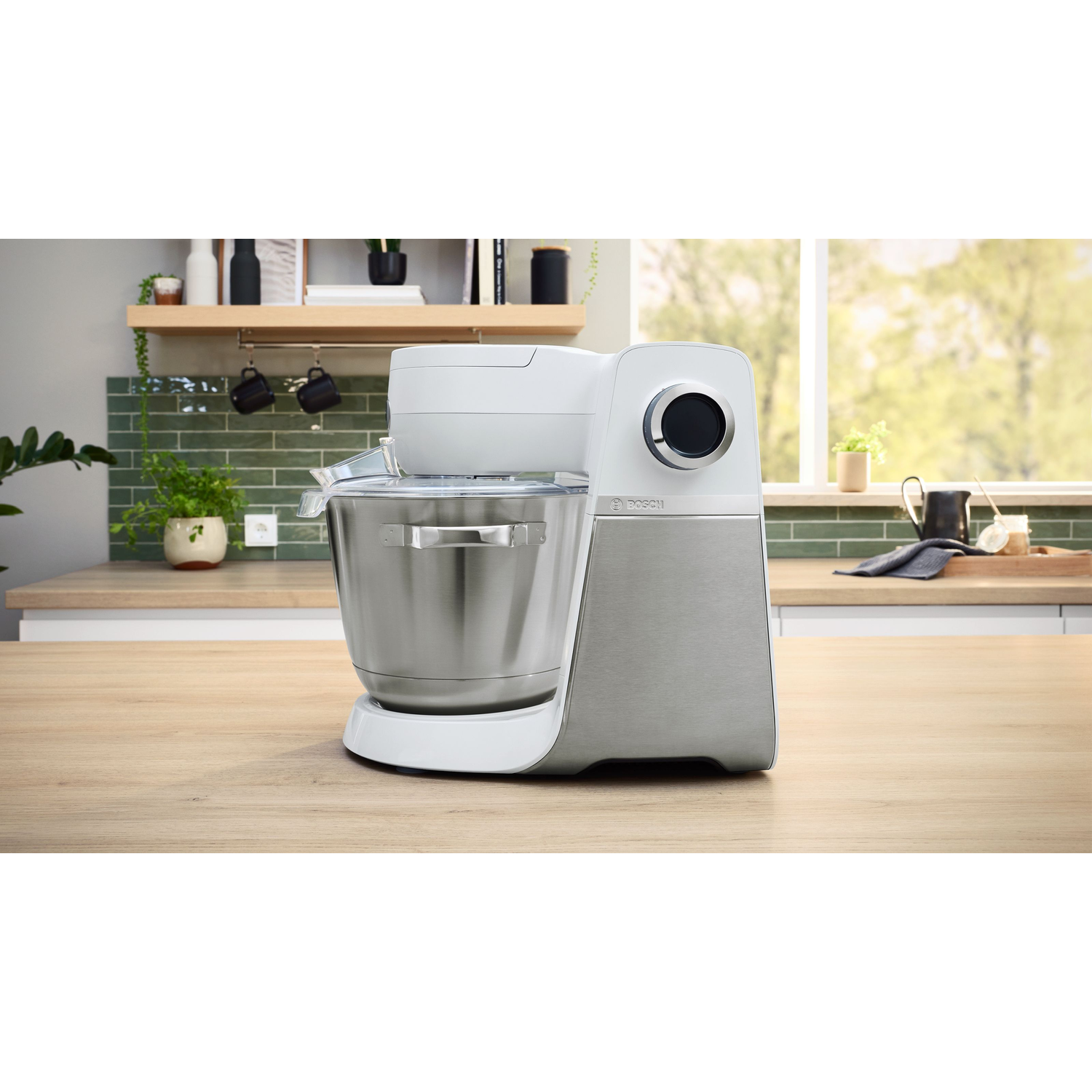A white and silver stand mixer on a wooden counter. Background features a kitchen with plants, a window, and shelves.