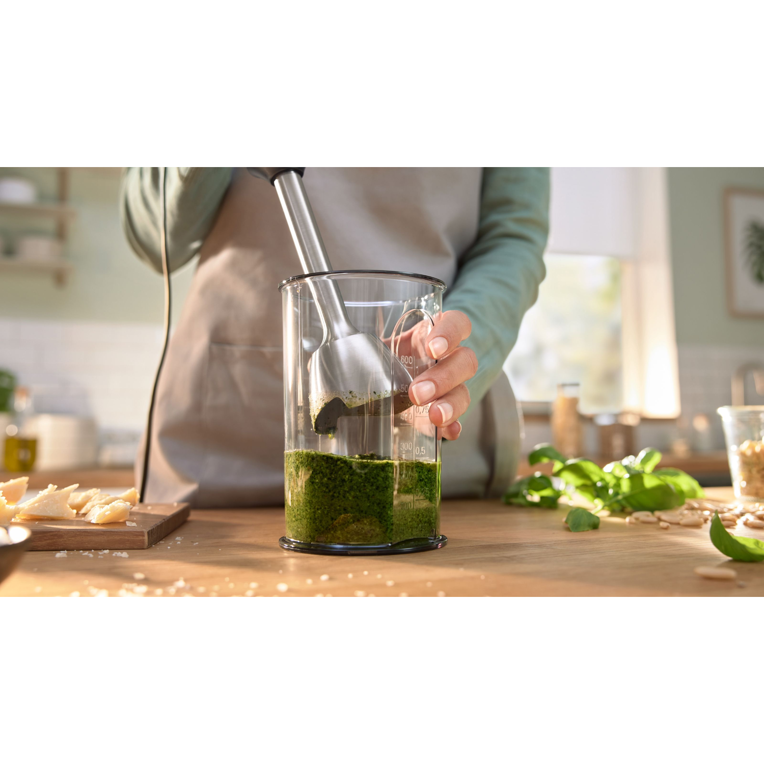 Een persoon mixt pesto met een staafmixer in een glas. Ingrediënten zoals basilicum, pijnboompitten en Parmezaanse kaas liggen op tafel.