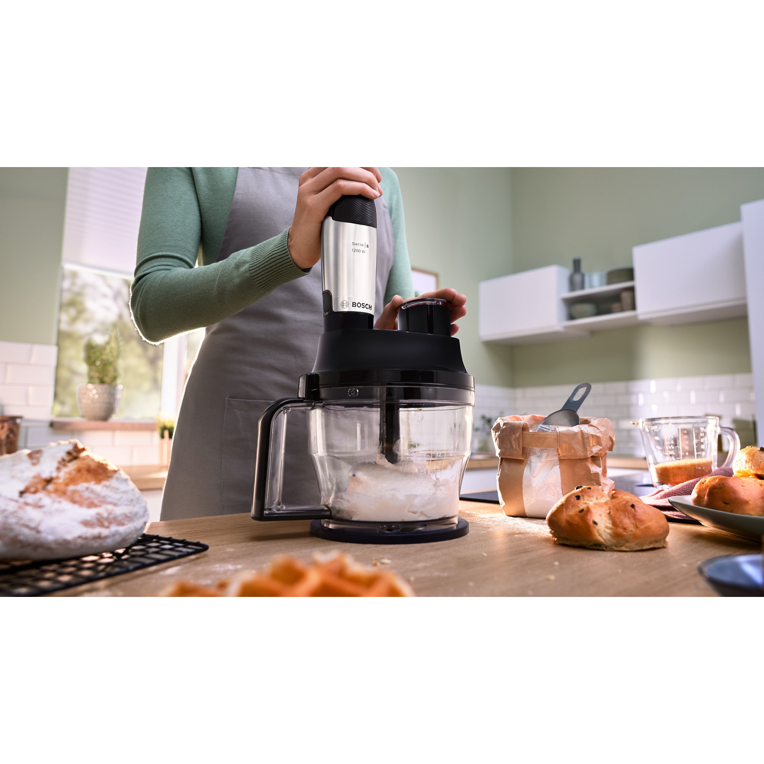 A person uses a food processor with dough inside. Bread, flour, and other ingredients are on the wooden countertop.