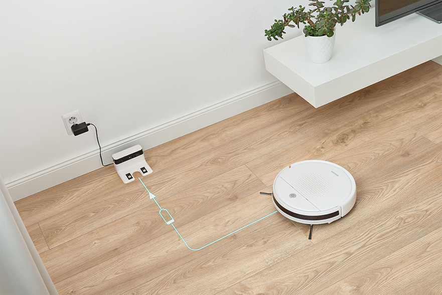 Fehér robotporszívó töltődik a fa padlón. Töltőállomás és fali aljzat.