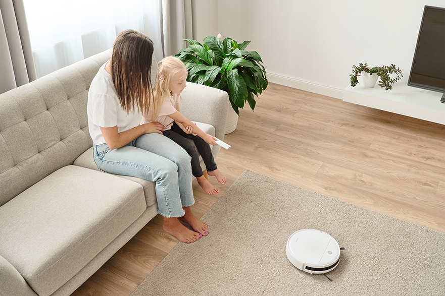 Egy nő és egy gyermek a kanapén egy robotporszívót néznek a szőnyegen. Szoba növényekkel.