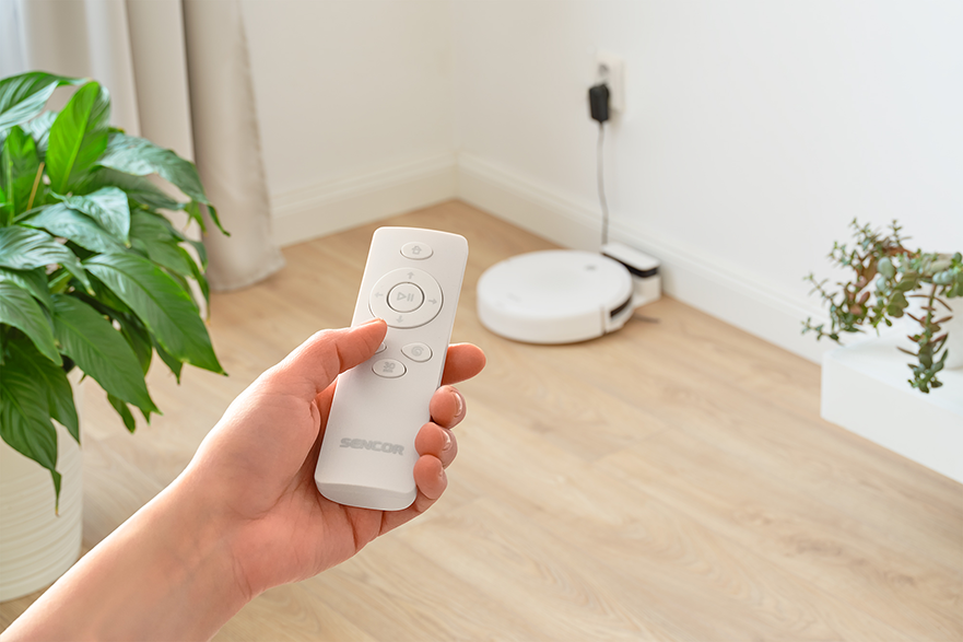 Egy kéz fehér távirányítót tart, a háttérben robotporszívó.