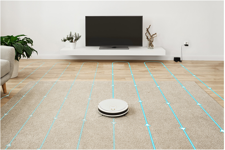 Robot porszívó szőnyegen, takarít, kék útvonalvonalakkal. TV és bútorok a háttérben.