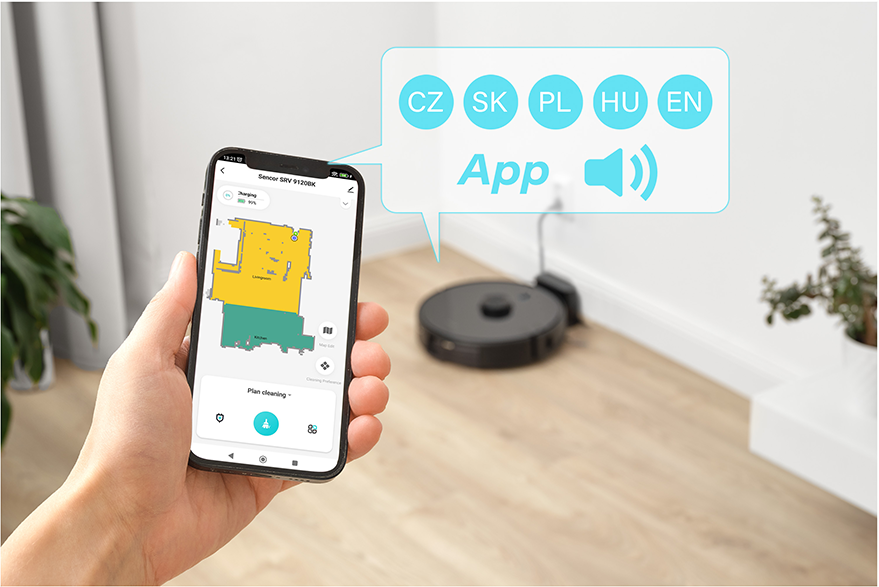 Egy kéz, amely egy telefont tart, rajta egy takarítási térképpel, a háttérben egy robotporszívóval.