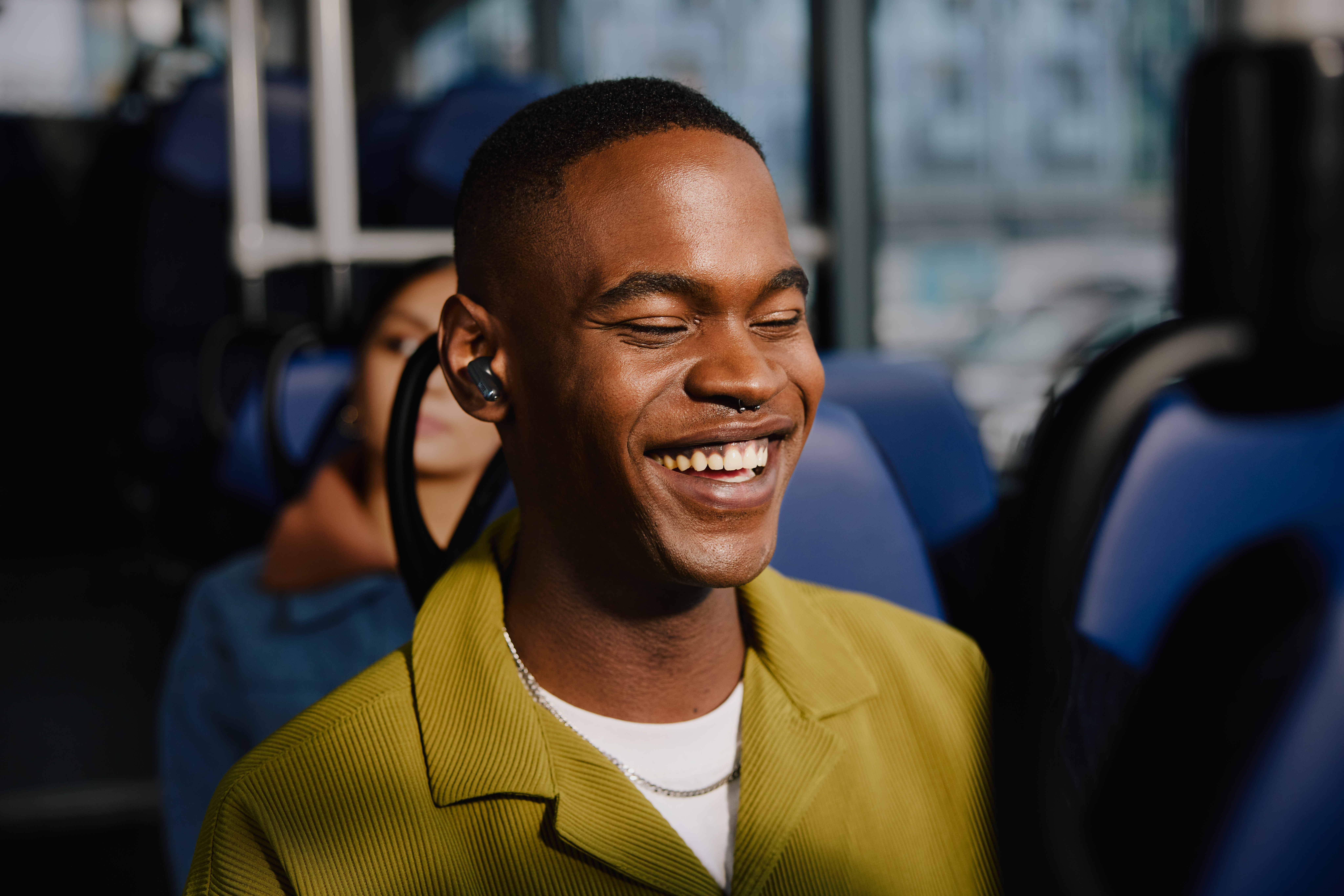 Ein lächelnder Schwarzer Mann mit Ohrhörern in einem Bus, eine weitere Person im Hintergrund.