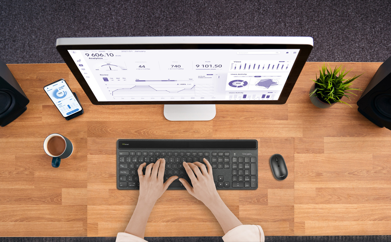 Persona escribiendo en un teclado, con un monitor mostrando datos, teléfono y planta.