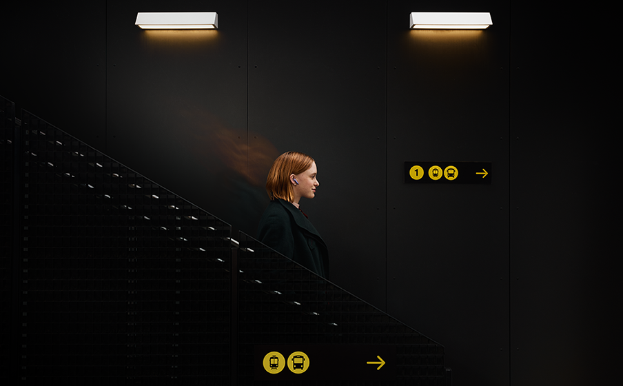 Une femme en manteau sombre descend les escaliers sous deux lumières et un panneau.