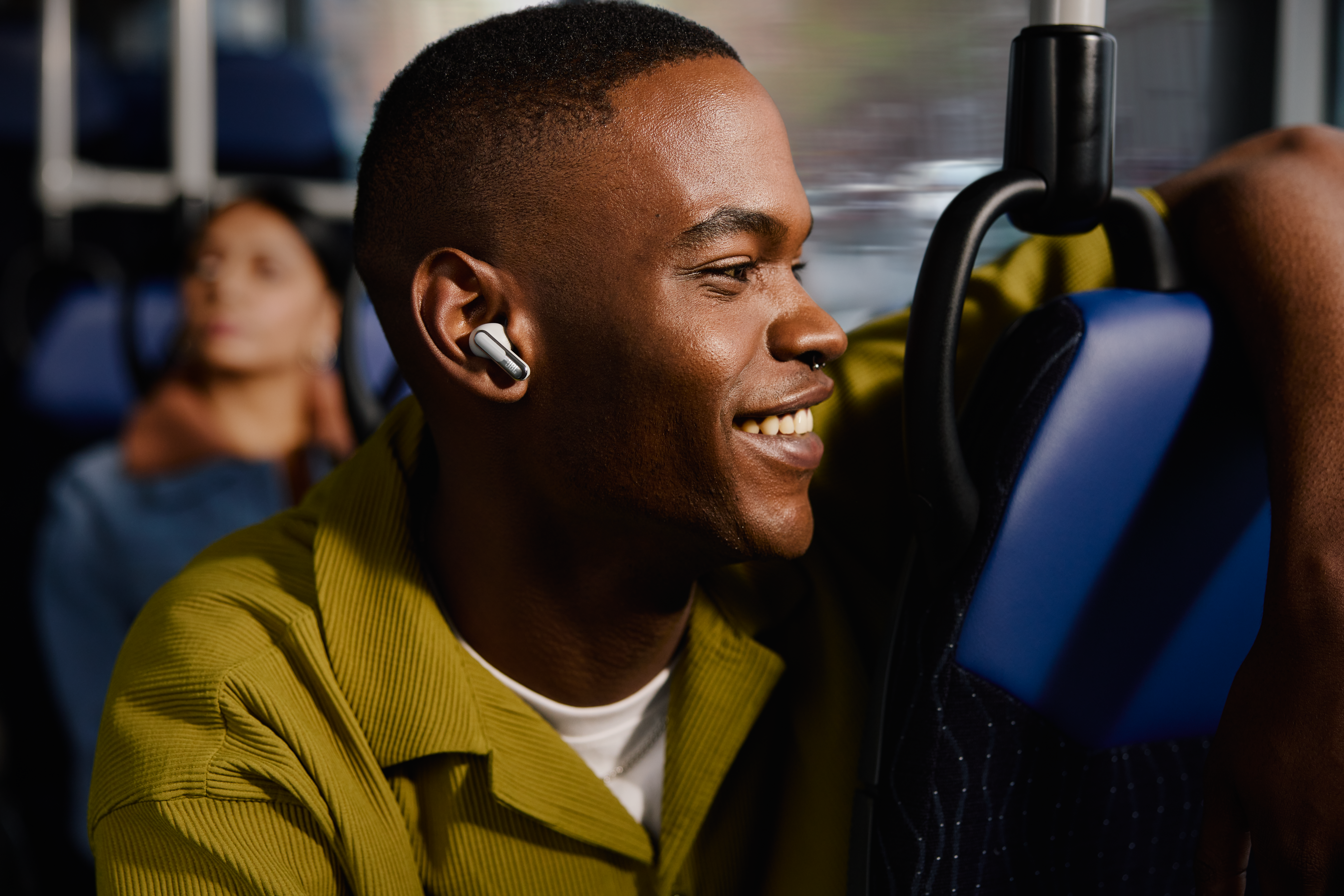 Un homme souriant en chemise verte portant des écouteurs, assis dans un bus. Une autre personne est en arrière-plan.