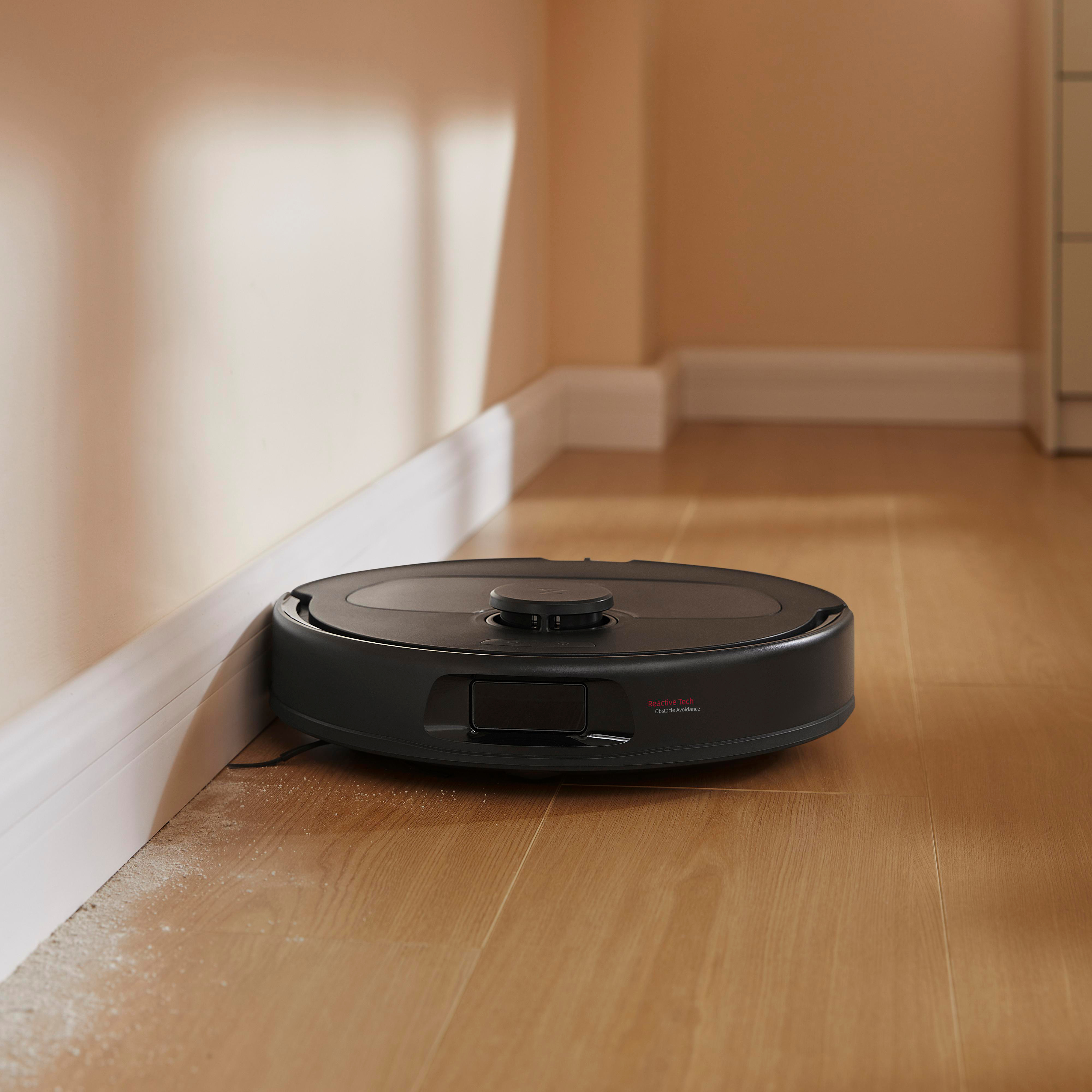 Ein schwarzer Roboter-Staubsauger reinigt einen Raum. Er befindet sich in der Nähe einer Wand und einer Fußleiste, mit etwas Schmutz.
