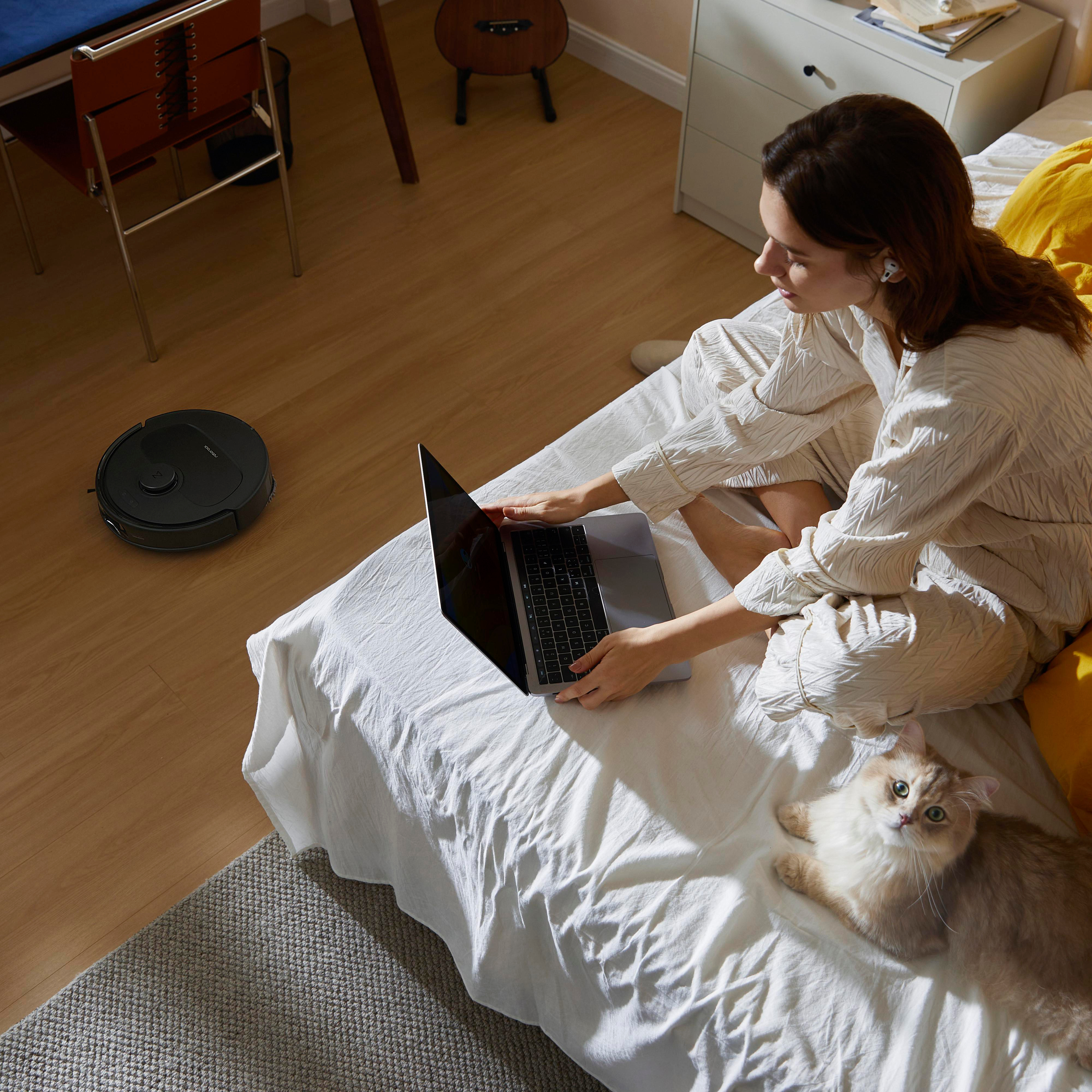 Eine Frau sitzt mit einem Laptop und einer Katze auf einem Bett. Ein Roboterstaubsauger ist im Bild.