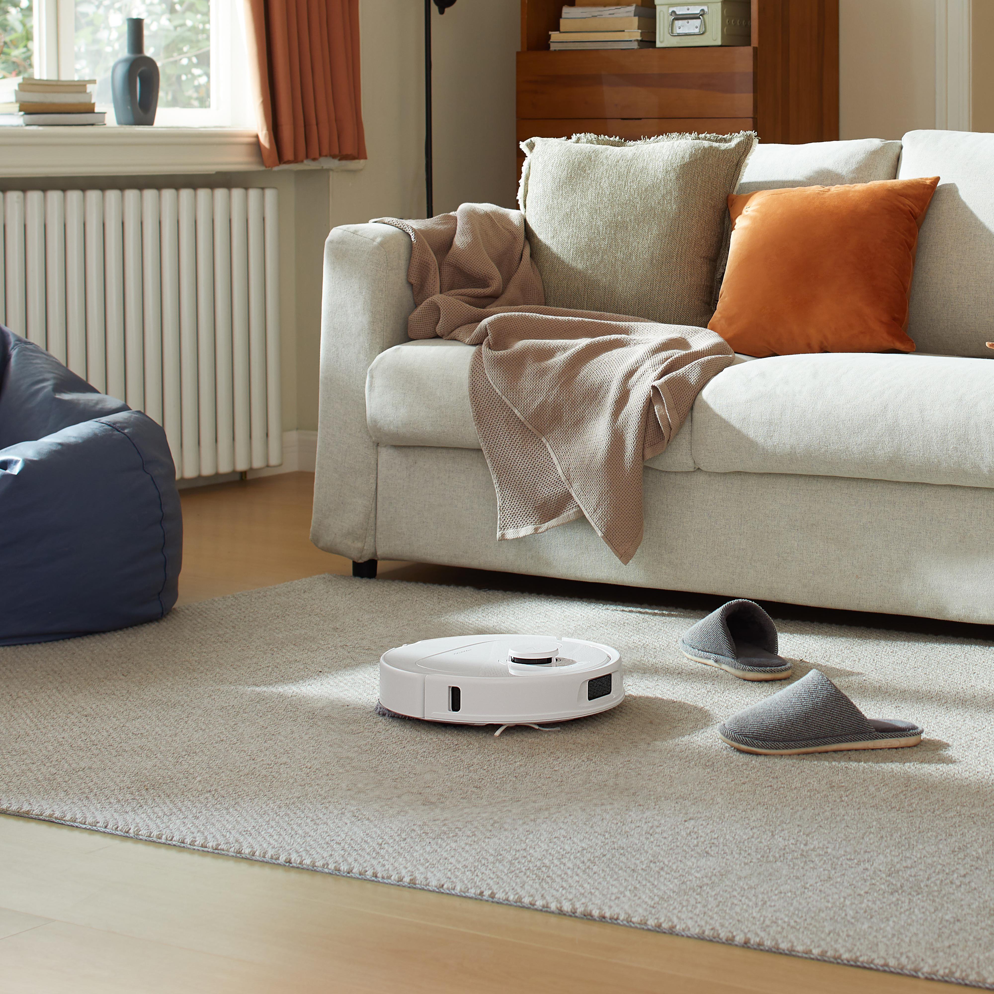 Ein weißer Roboter-Staubsauger auf hellem Teppich mit Hausschuhen und Sofa.