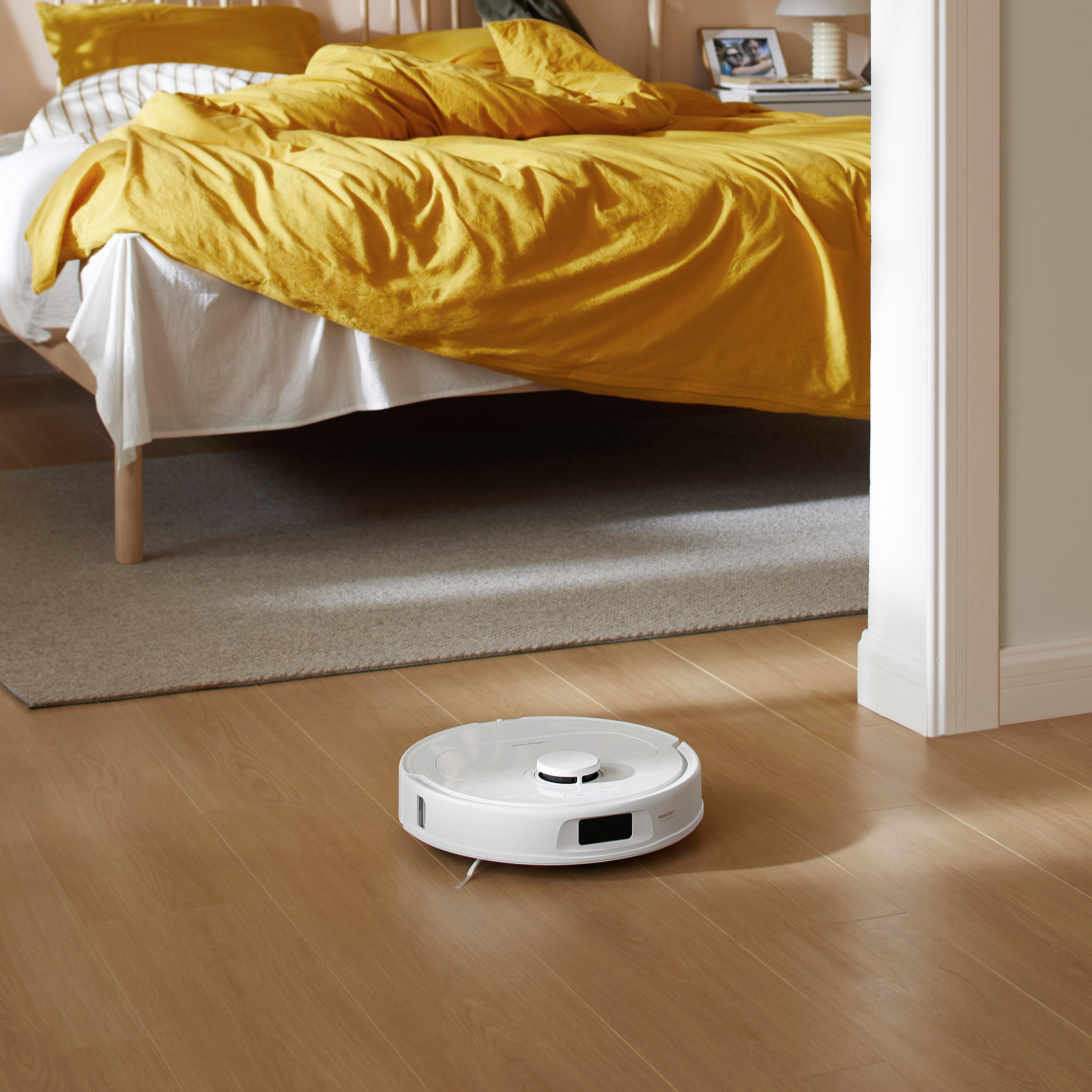 Roboter-Staubsauger auf Holzboden. Ein Bett mit gelben Bezügen ist im Hintergrund.
