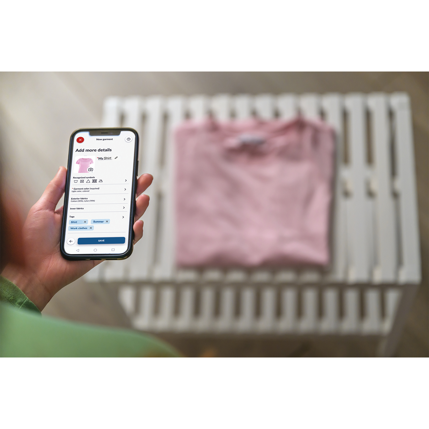 Una persona tiene in mano un telefono con un'app, accanto a una maglietta rosa su un supporto bianco. Lo sfondo è sfocato.